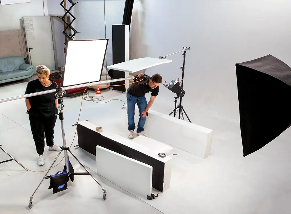 Dreipersonen in einem Fotostudio bei der Produktion eines Fotos, mit Licht- und Kameraausrüstung, weißen und schwarzen Platzhaltern, und einer grauen Hintergrundwand.