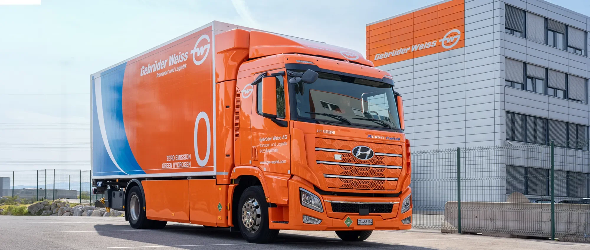 Ein orangefarbener Lastwagen der Firma Gebrüder Weiss liegt vor einem modernen Bürogebäude. Der Lastwagen ist elektrisch und wird mit Wasserstoff betrieben, was durch die Aufschrift "Zero Emission Green Hydrogen" auf der Seite angezeigt wird.