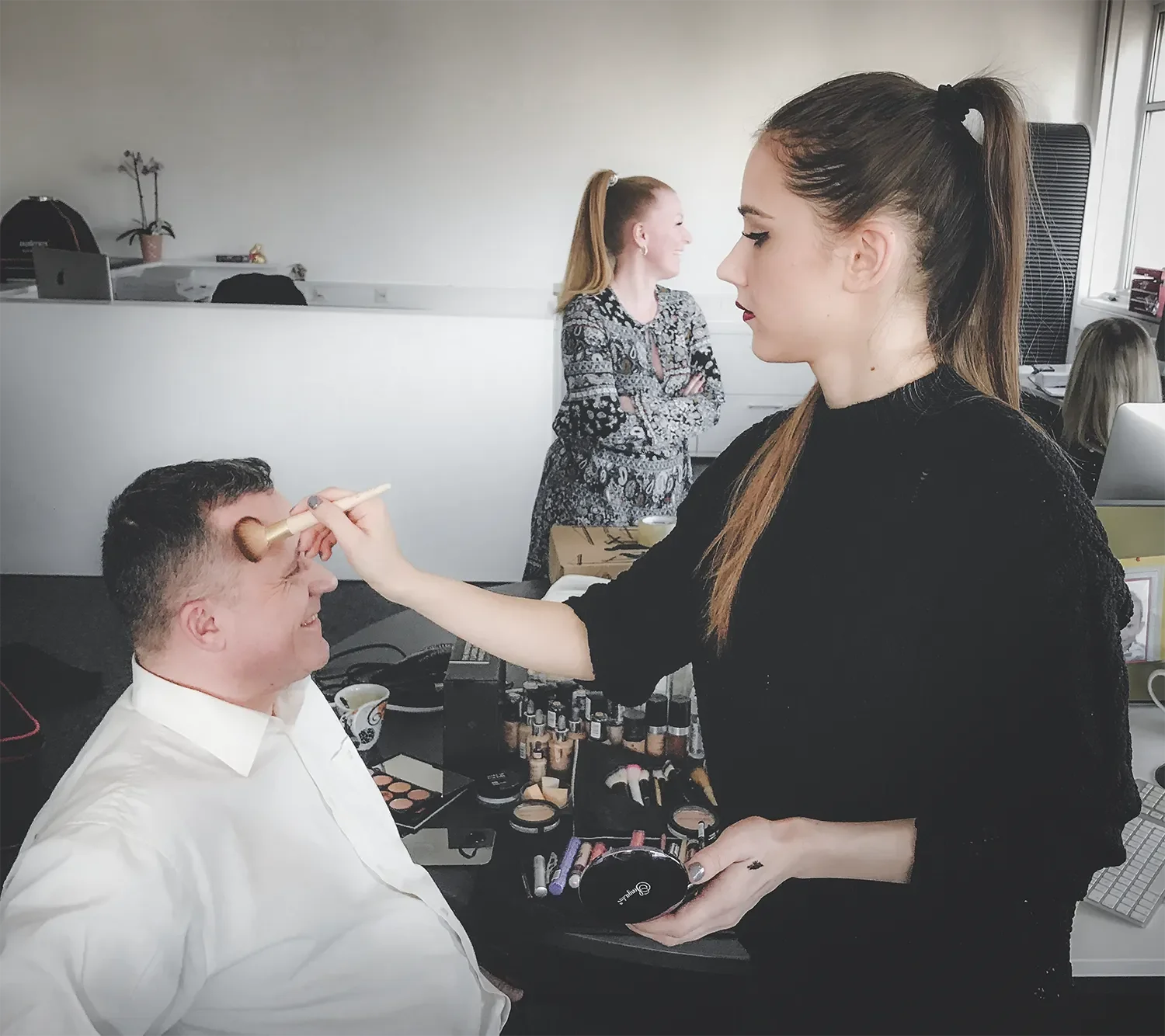 Frau trägt Make-up bei einem Mann, im Hintergrund eine weitere Frau, die lächelt, in einem Büro oder Studio mit Make-up-Produkten auf einem Tisch.