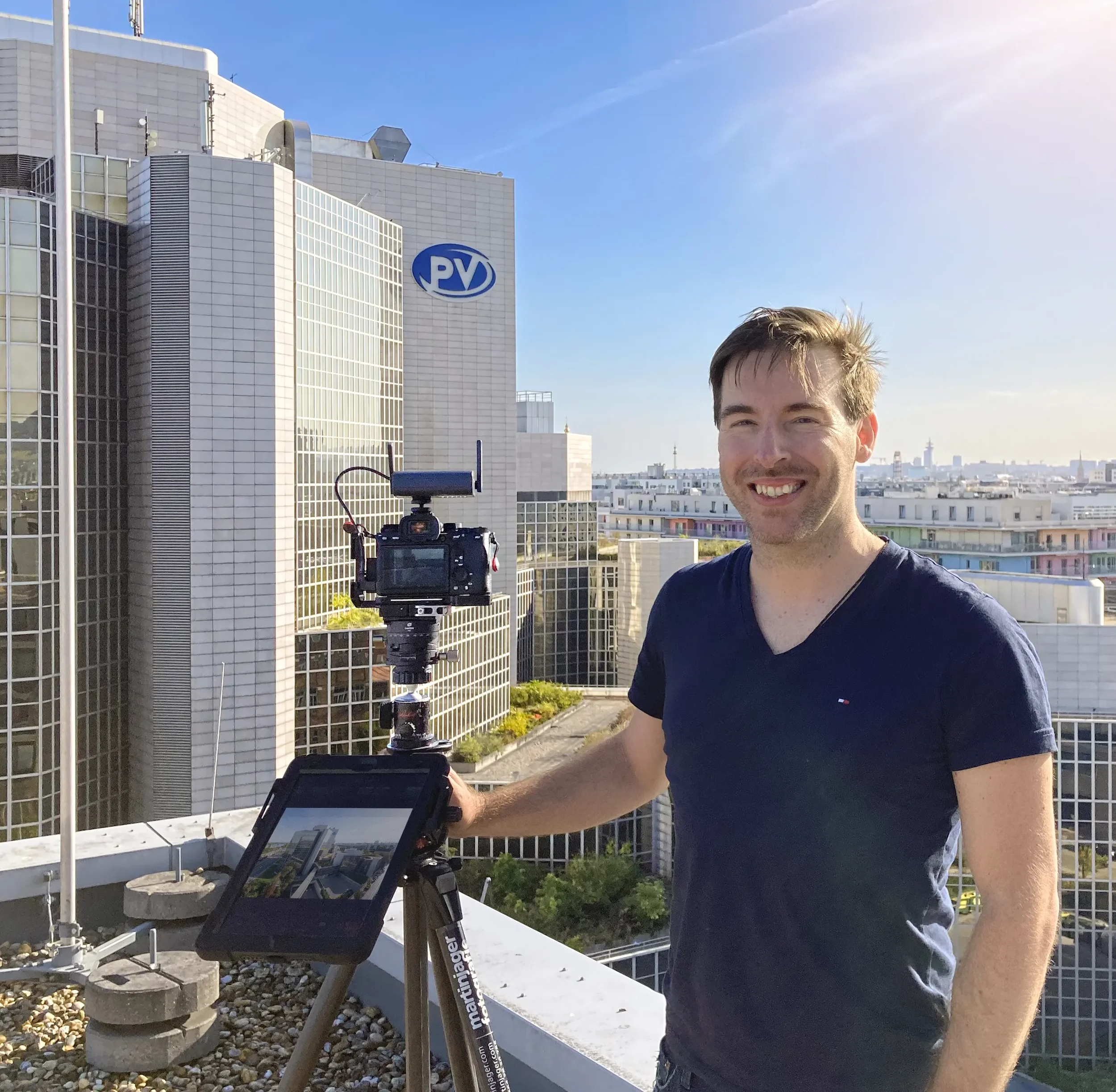 Junger Mann auf einem Balkon mit Hochhaus im Hintergrund, steht vor einem Stativ mit Kamera, im Sonnenlicht, blauer Himmel.