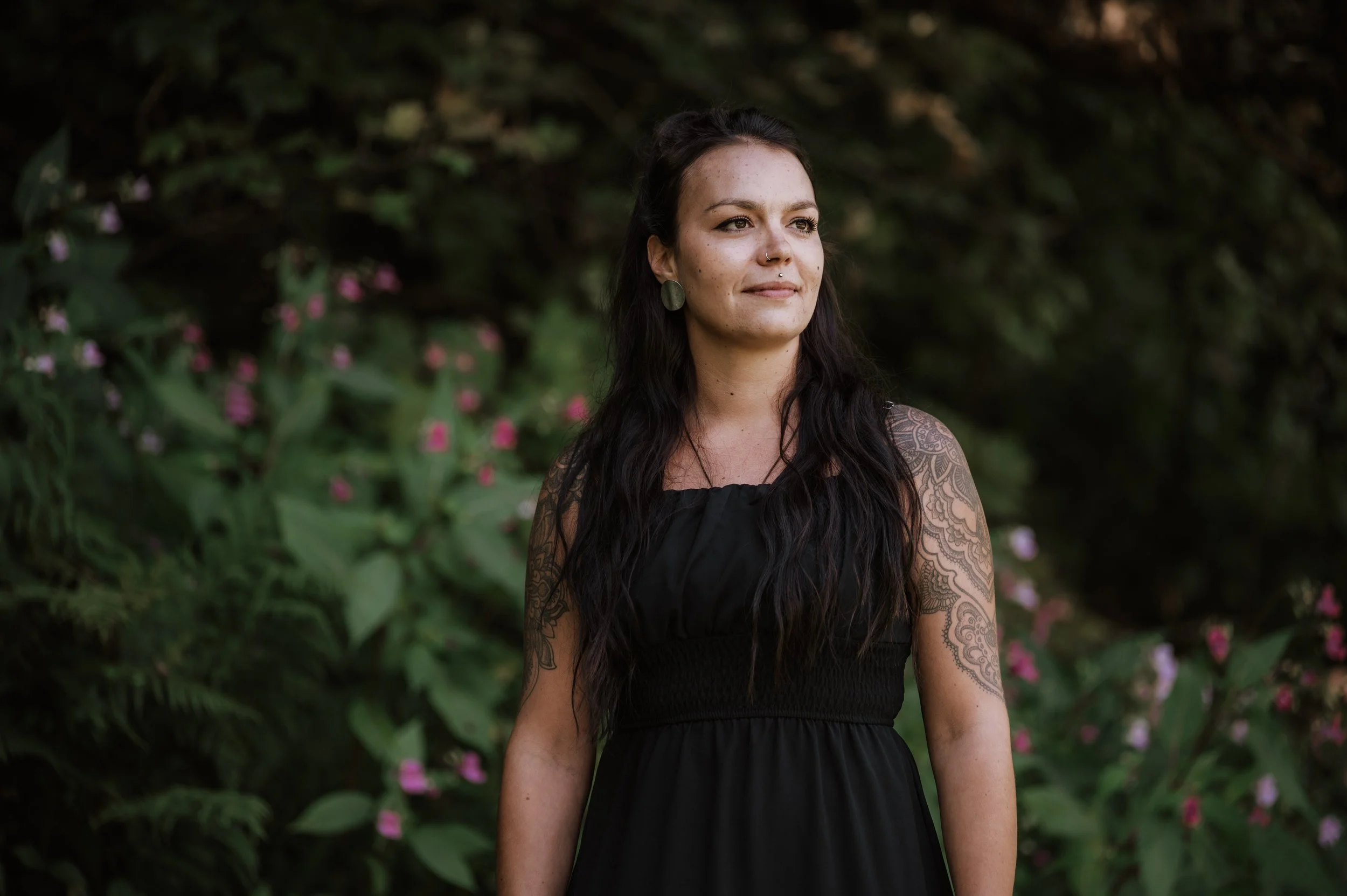 Junge Frau mit Tattoos trägt schwarzes Kleid, steht vor grüner Pflanzenwand mit pinken Blumen.