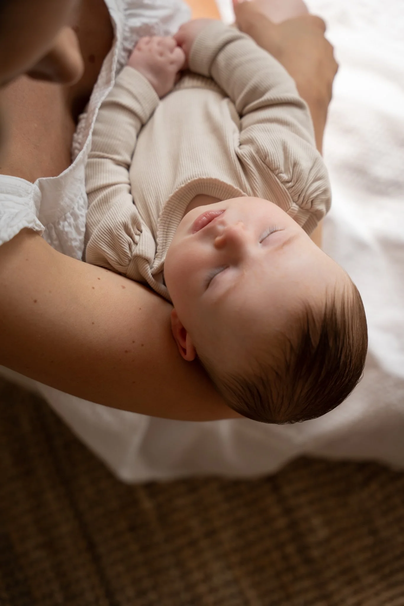 Een slapende baby wordt vastgehouden door een persoon, mogelijk de moeder, met de baby op haar arm terechtgekomen. De baby draagt een beige outfit en ligt comfortabel met gesloten ogen.