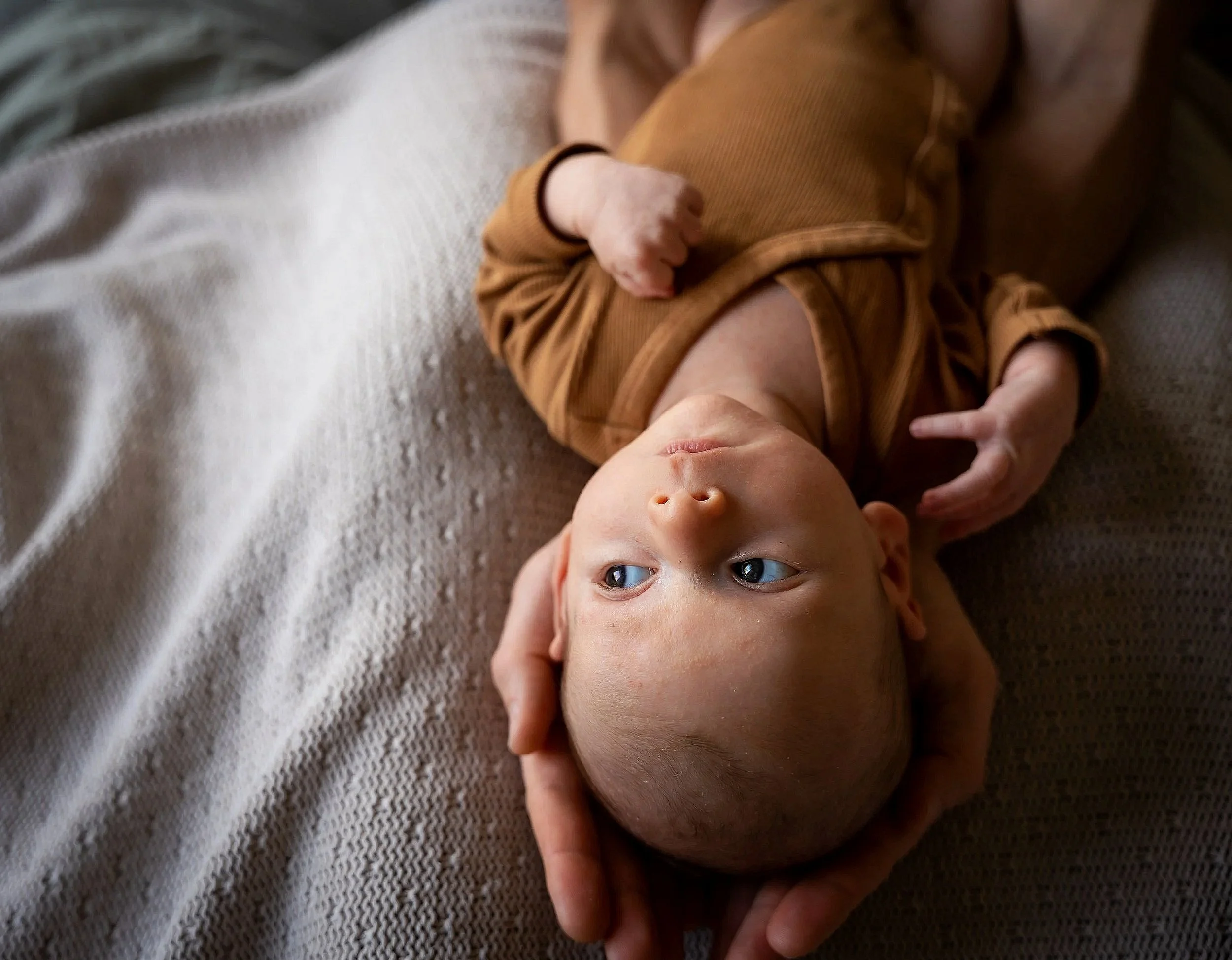 Newborn fotografie aan huis in Terheijden. Baby ligt op een grijze deken, gekleed in een bruin outfit, en wordt zachtjes vastgehouden door iemands handen.