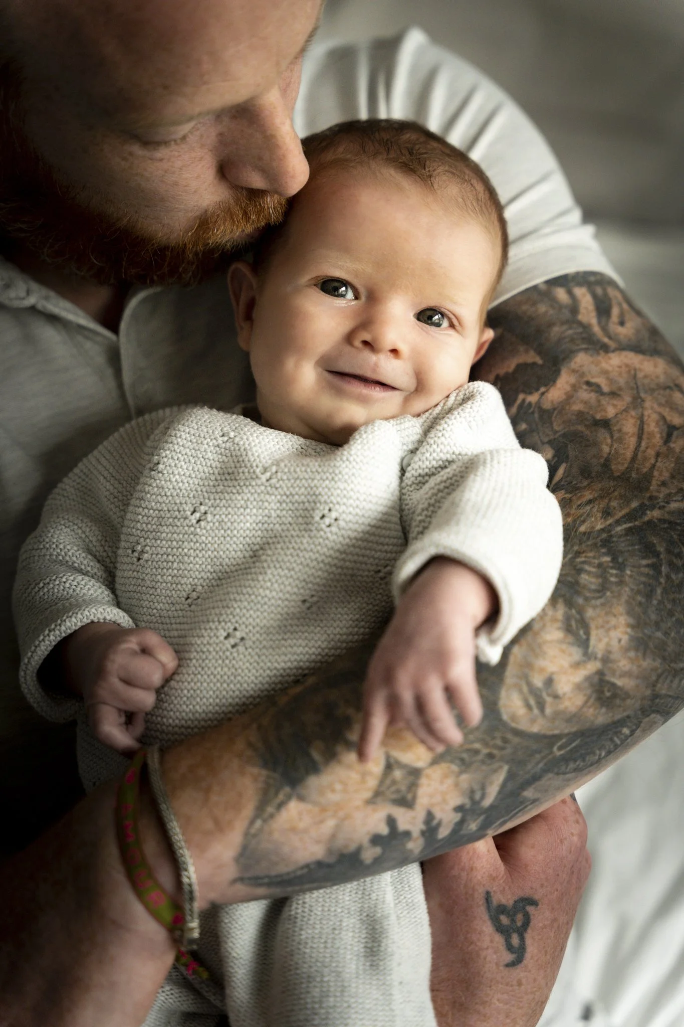 Een man met tatoeages en een lichte huid houdt een lachend babymeisje met blauwe ogen en rood haar vast.