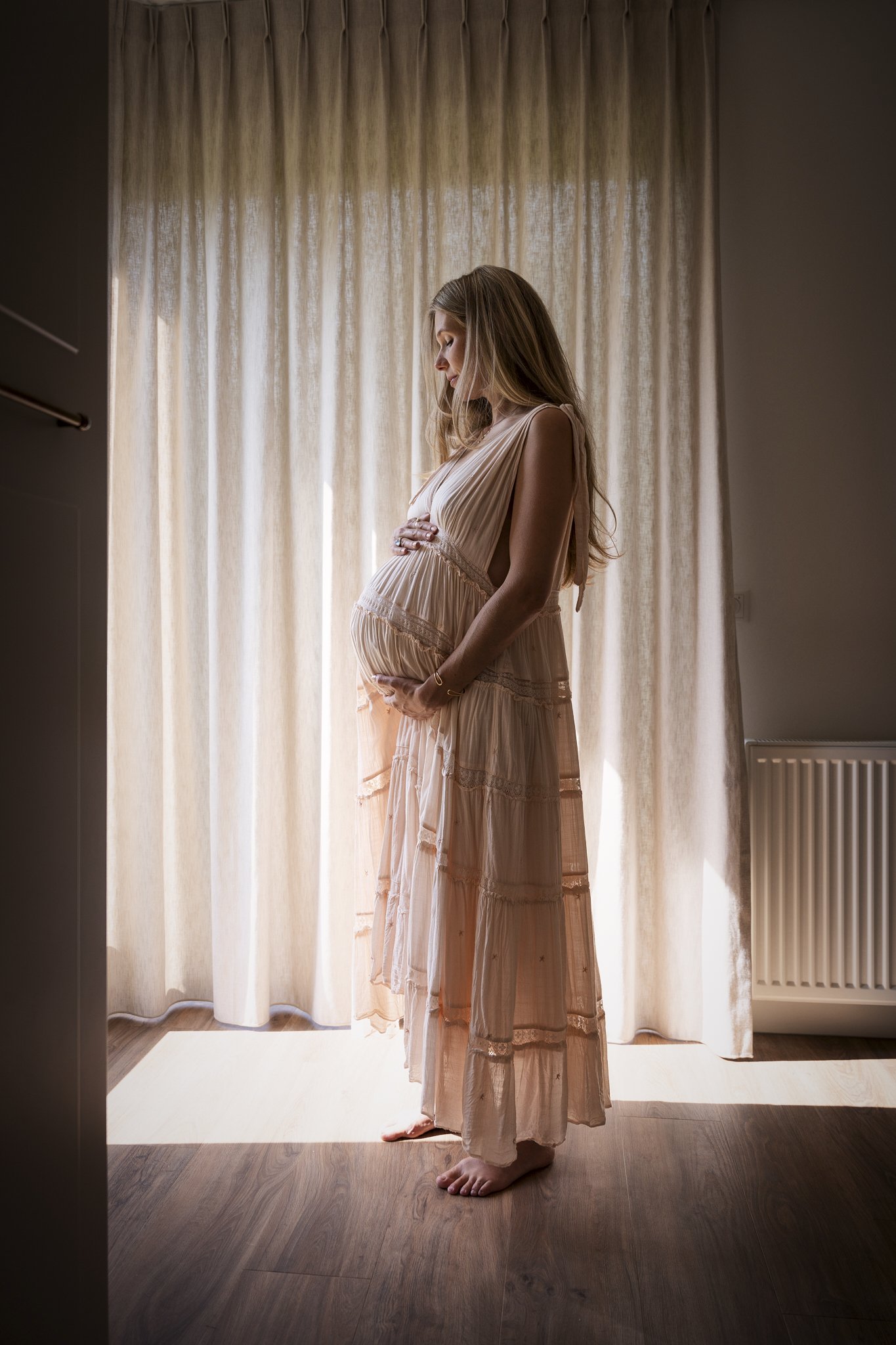 Zwangere vrouw in zijaanzicht die haar buik vasthoudt, binnen bij daglicht, zachte pastelkleuren. Gefotografeerd door zwangerschapsfotograaf.