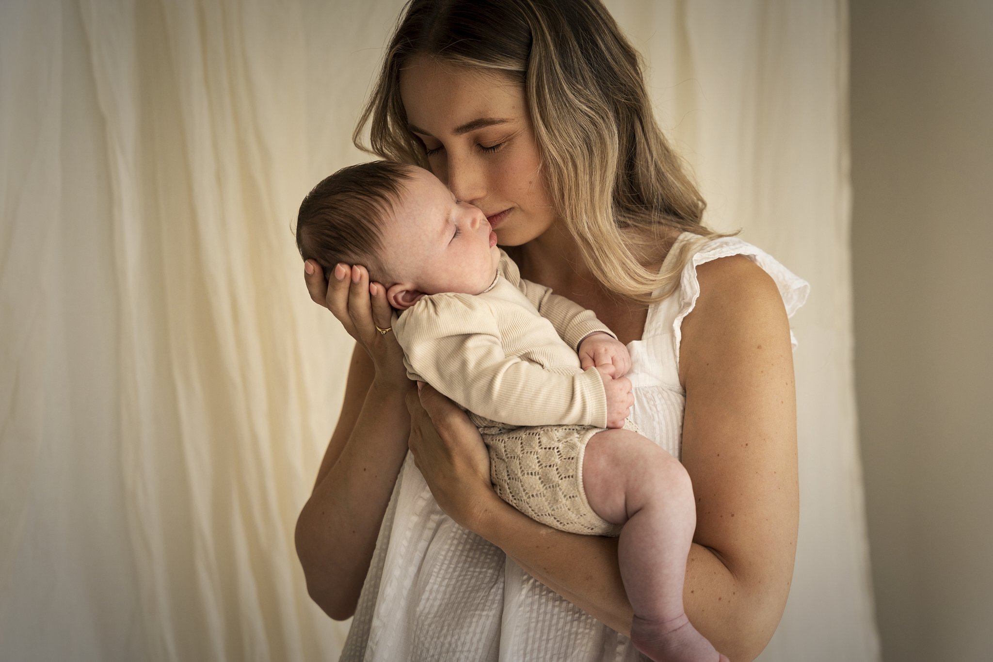Moeder houdt baby dicht tegen haar gezicht, beide met gesloten ogen, binnen, met lichte gordijnen op de achtergrond.