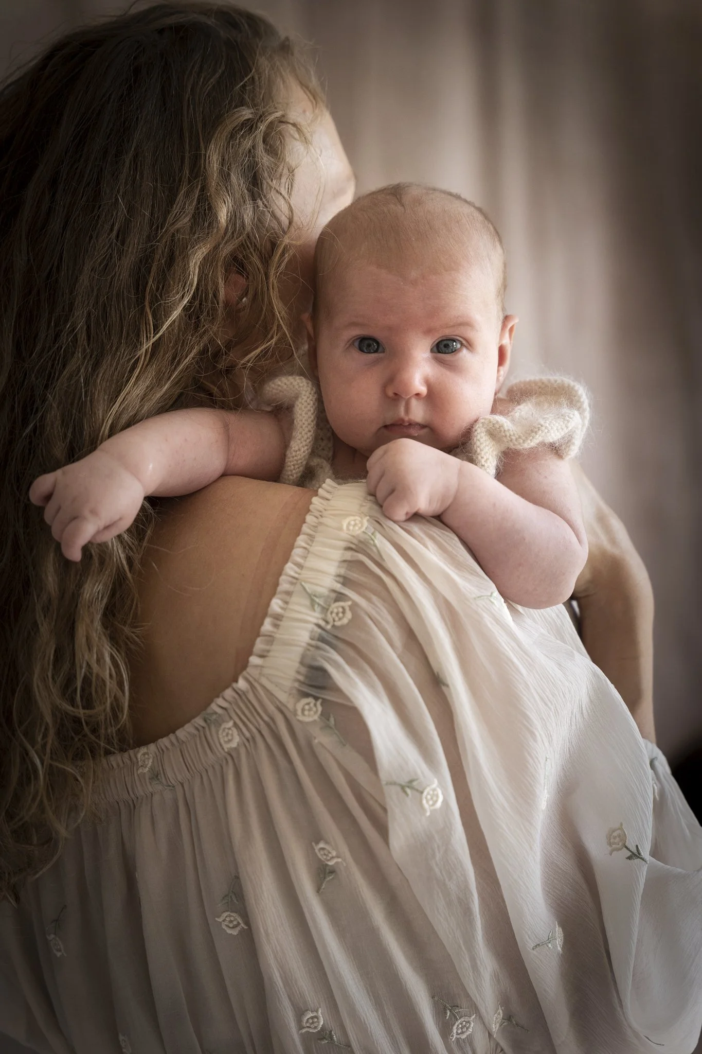 Close-up van een baby die op de schouder van een vrouw ligt, met de blik naar de camera gericht, in een interieur met zachte verlichting.