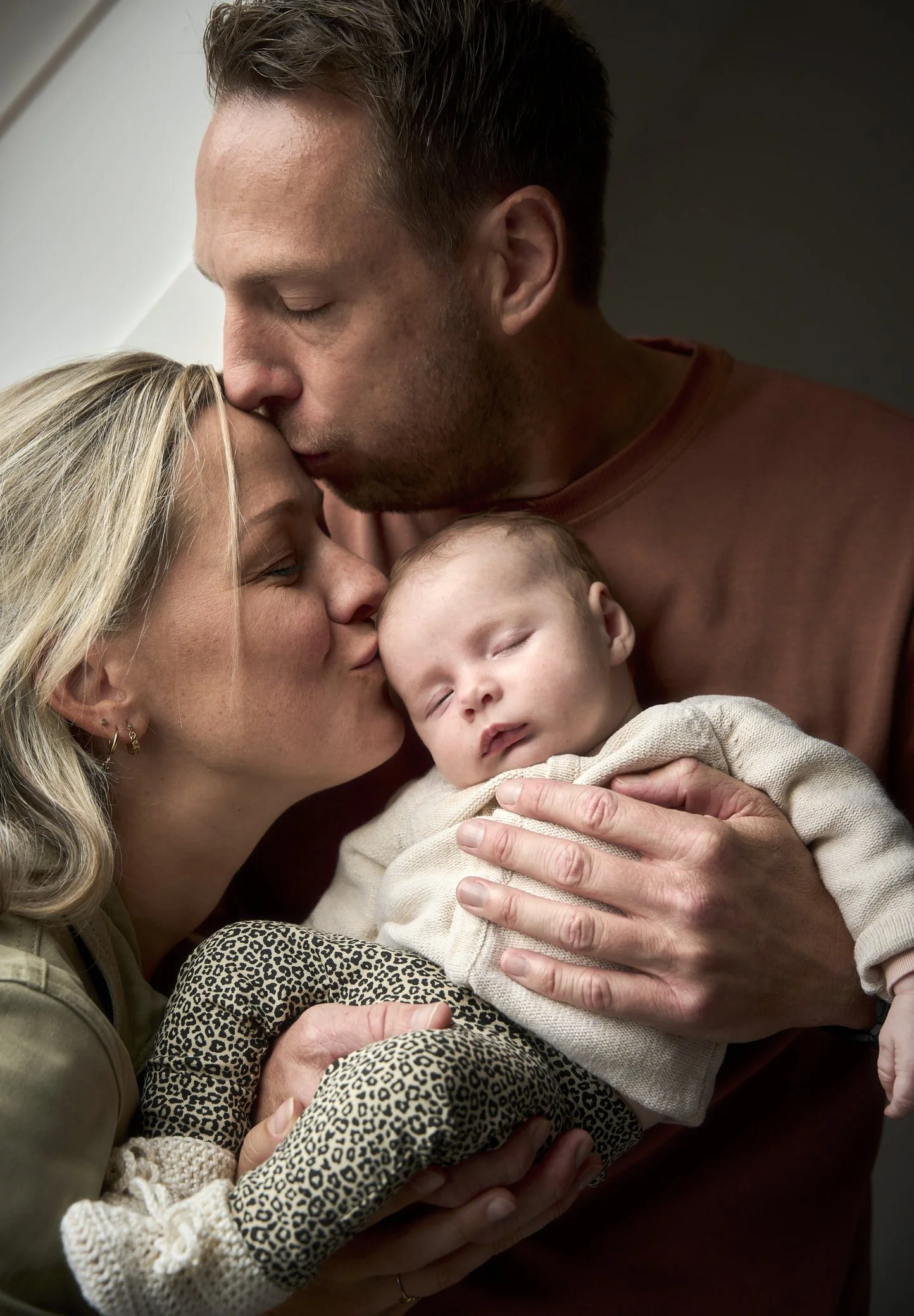 Een jong gezin bestaat uit een moeder, vader en hun slapende baby, waarbij de vader de baby vasthoudt en de moeder de baby kus.