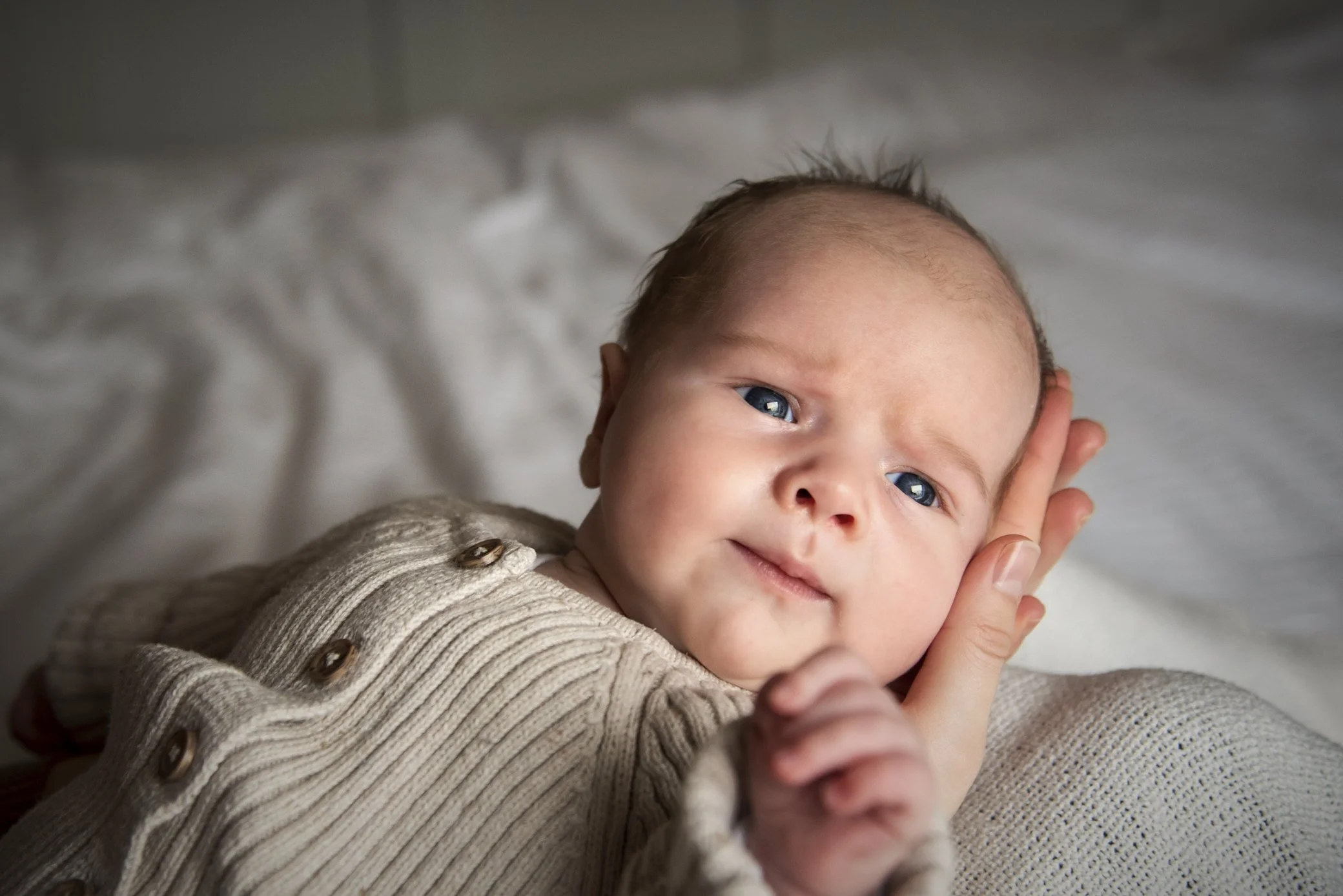 Een jonge baby ligt op een zachte witte deken, wordt zachtjes ondersteund door een hand, en kijkt lief naar de camera.