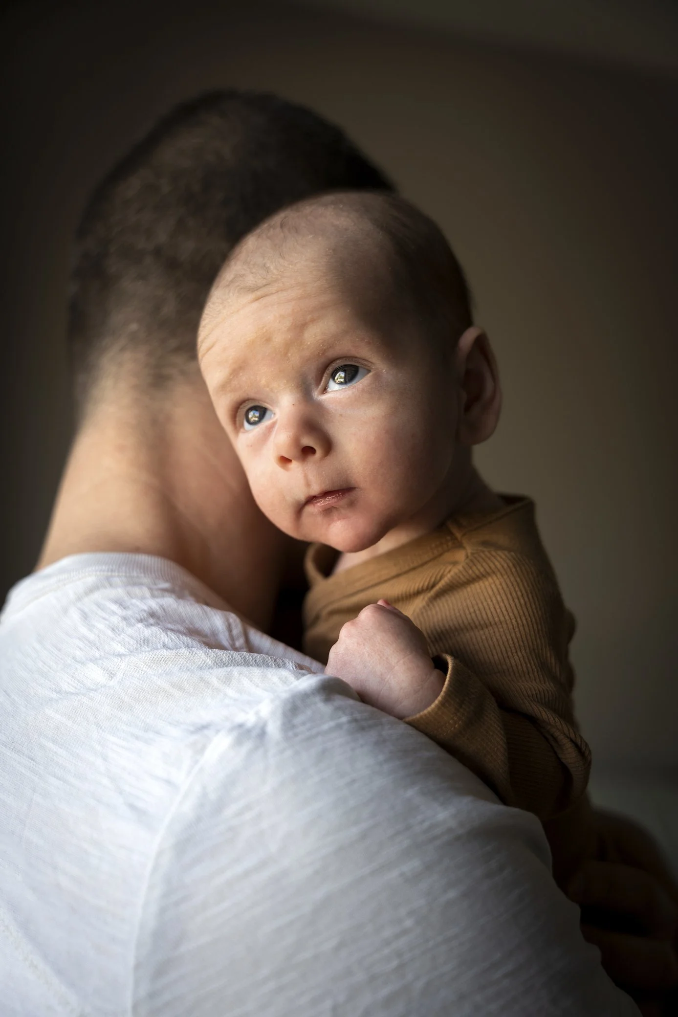 Newborn fotografie aan huis in Terheijden. Close-up van een baby die op de schouder van een volwassene rust, kijkt omhoog met een nieuwsgierige blik.