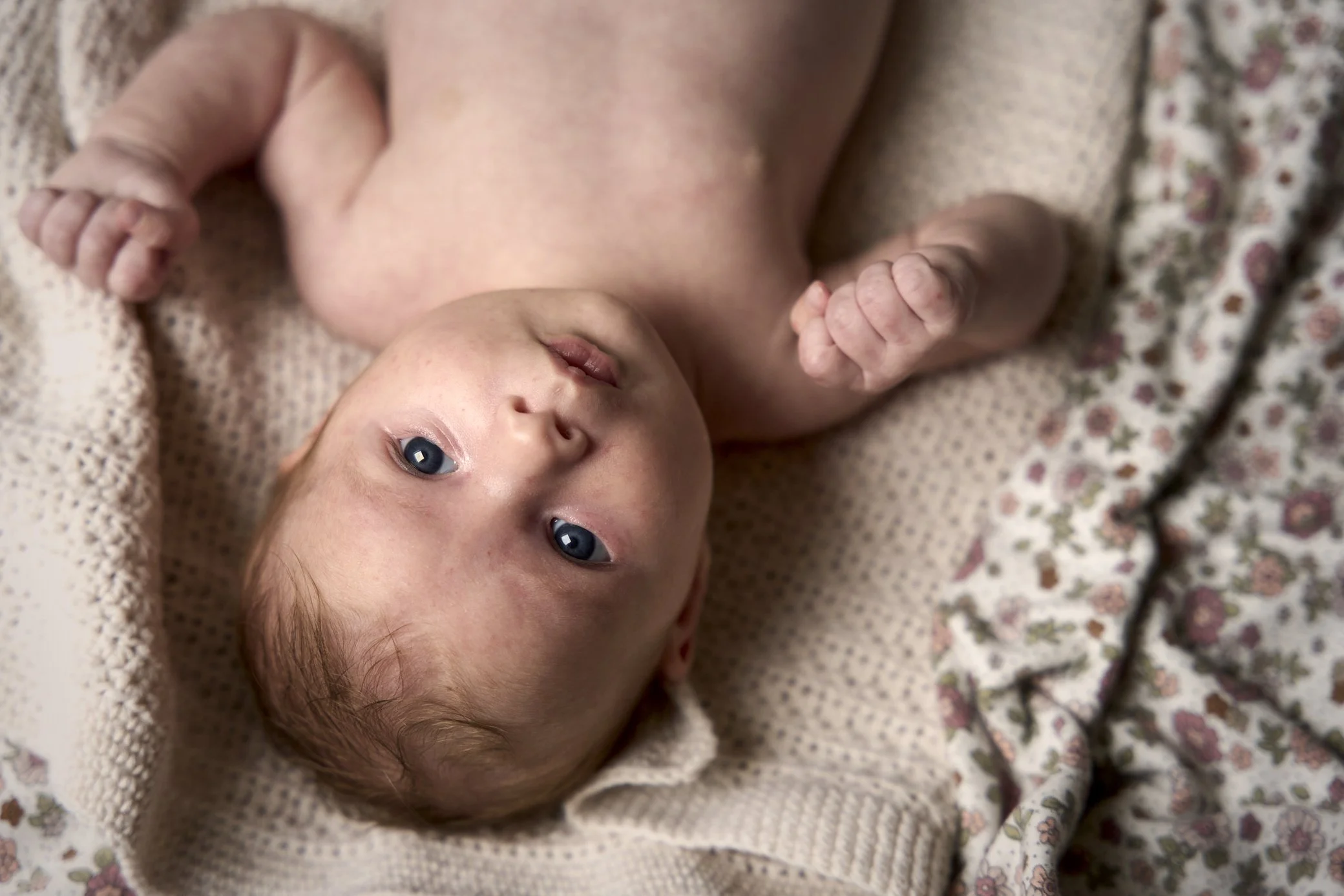 Babytje ligt op een gebreide deken, kijkt omhoog met blauwe ogen, heeft lichtbruin haar en is naakt.