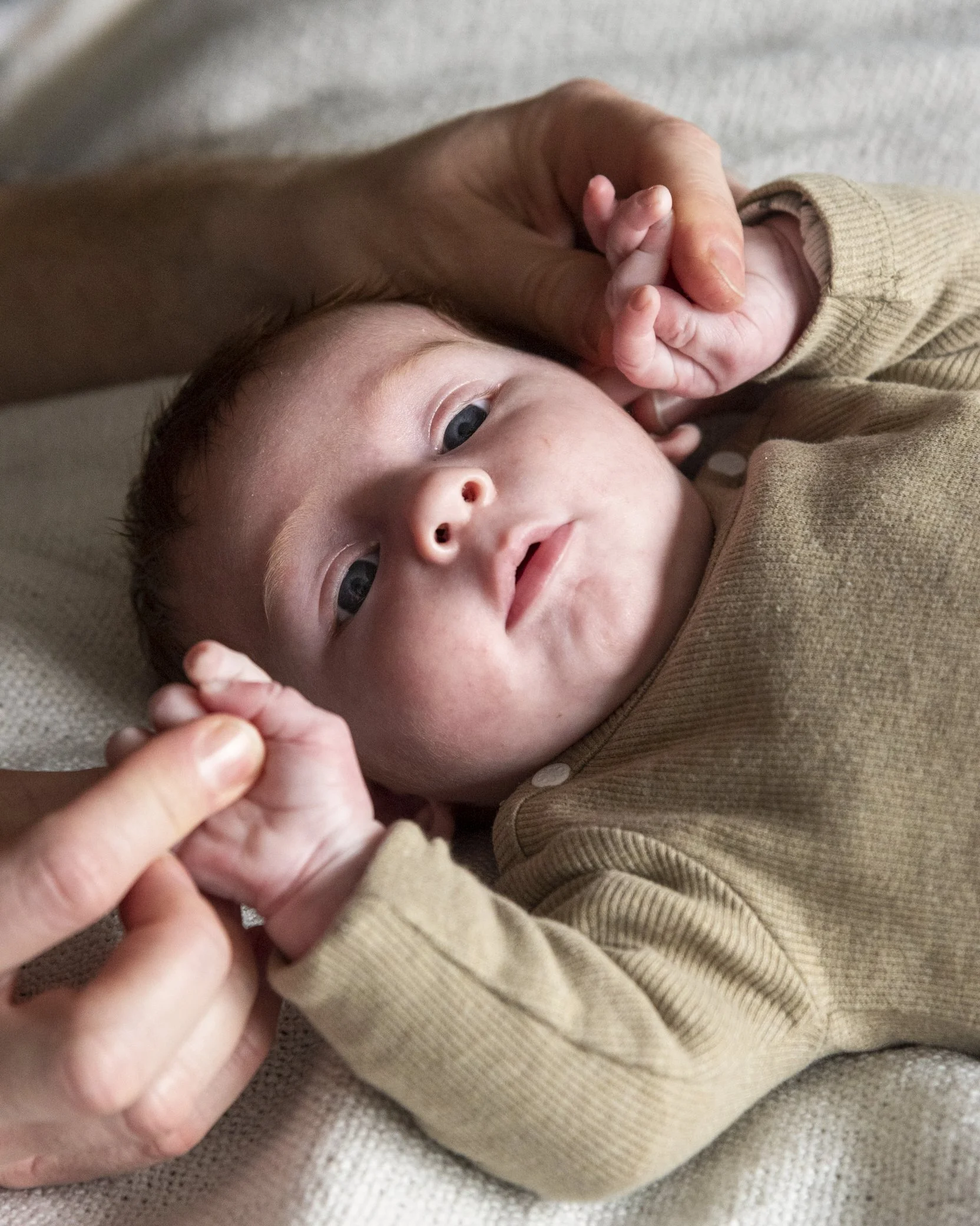 Een baby ligt op een zachte ondergrond, met een persoon die zachtjes een hand vasthoudt en een andere hand op het hoofd van de baby legt. De baby kijkt naar de camera met een ontspannen uitdrukking.