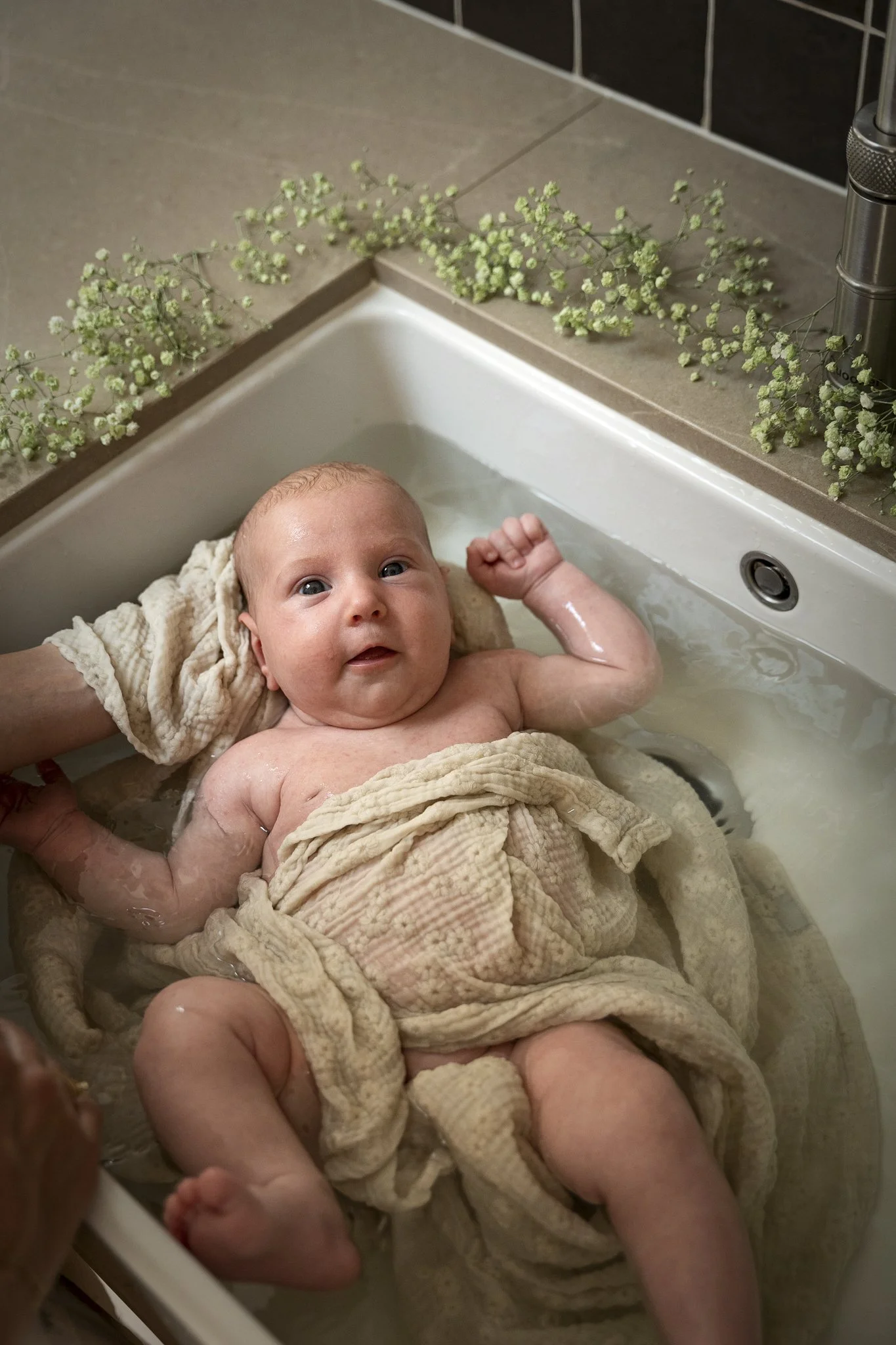 Een baby wordt in een badje verzorgd, omgeven door bloemen op de rand van de wastafel.