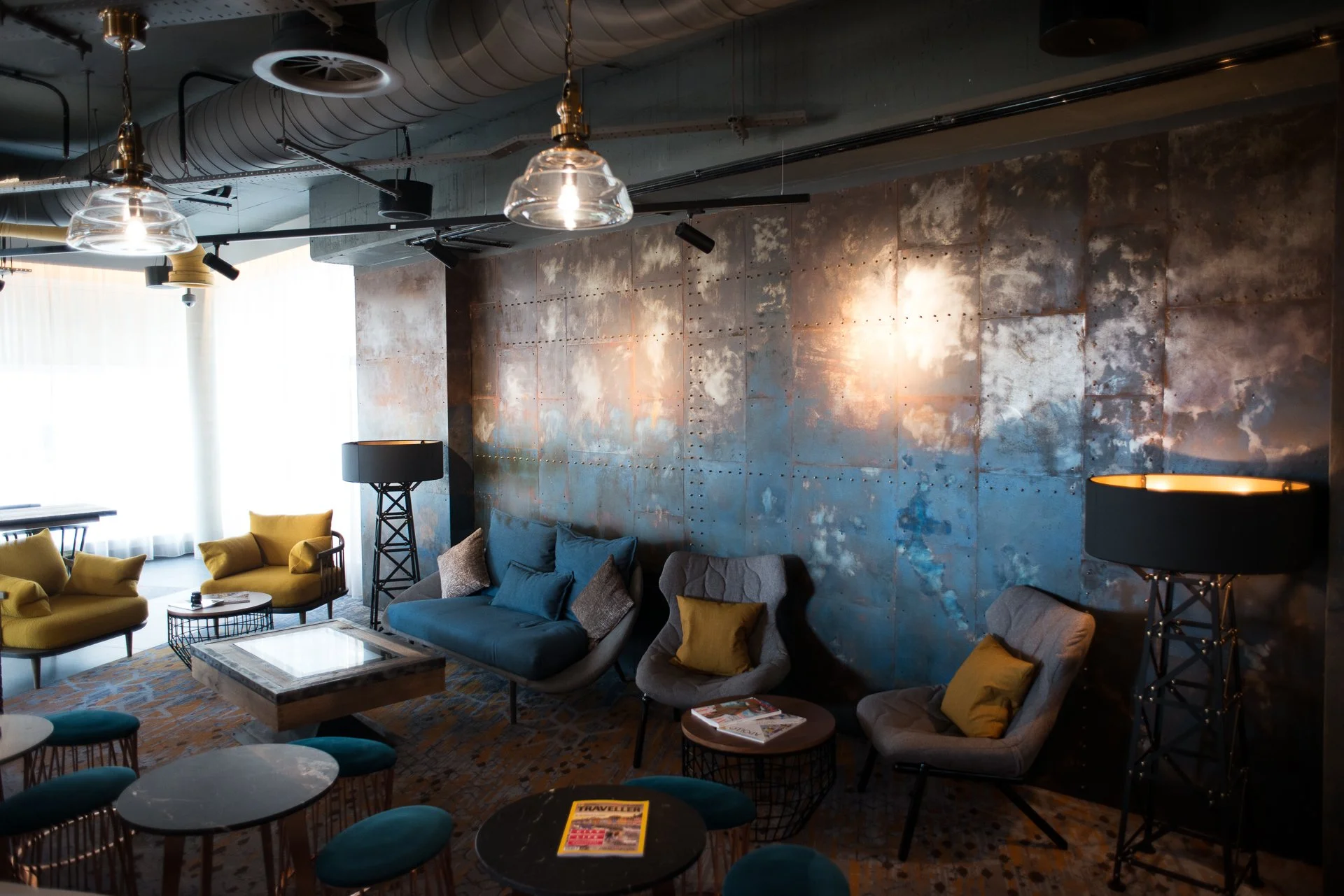 Modern living room with yellow and gray sofas, blue armchairs, a coffee table, and lamps, featuring an industrial-style ceiling with exposed pipes and a textured wall.