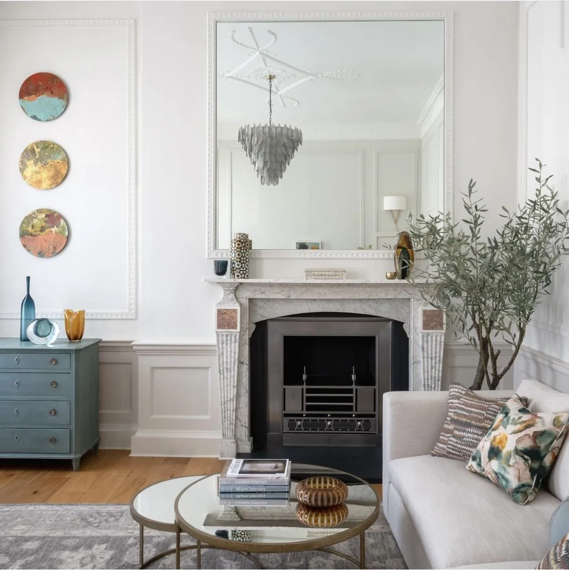 Living room with white walls, decorative wall art, a large mirror above a fireplace, a chandelier hanging from the ceiling, a green side table with vases, a large potted plant, a white sofa with colorful pillows, and a round coffee table with books a