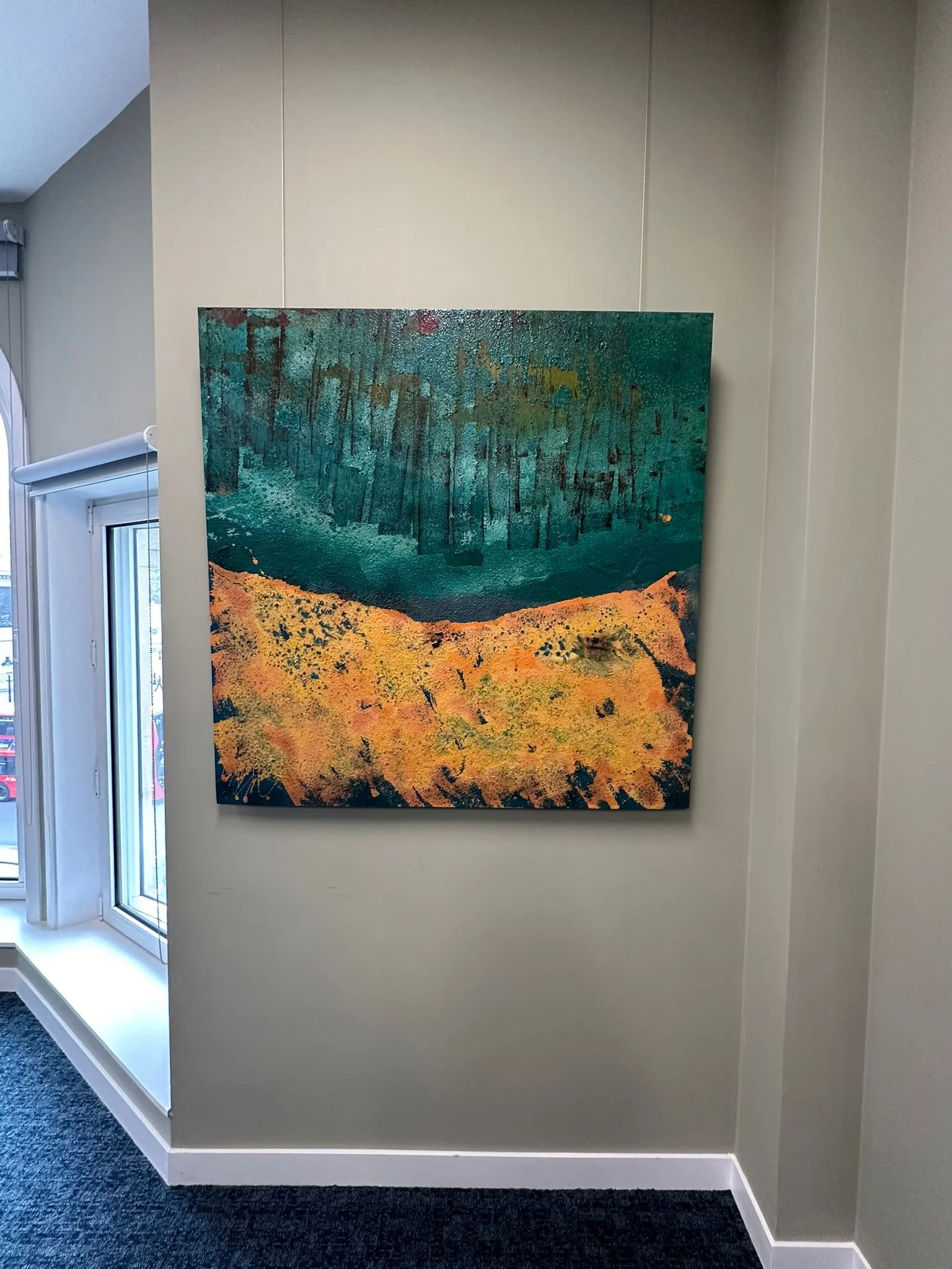 An abstract painting with blue, green, yellow, and orange colors hangs on a beige wall in a room with a window and a dark carpeted floor.