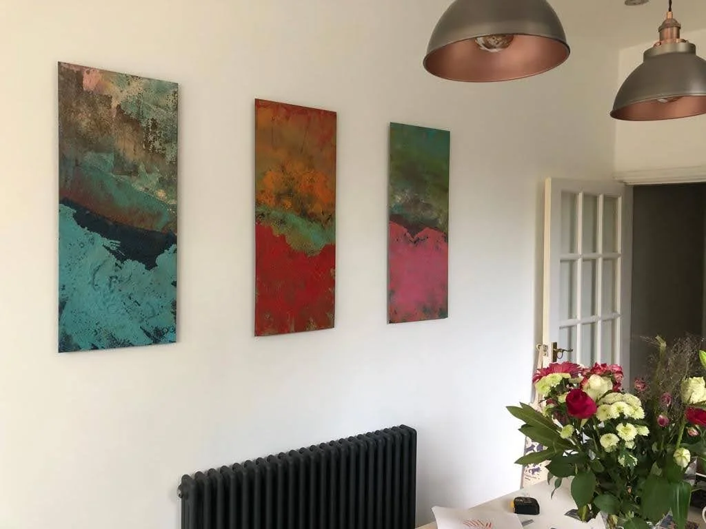 Three vertical abstract paintings on a white wall above a black radiator with a bouquet of mixed flowers and a remote control on a table.