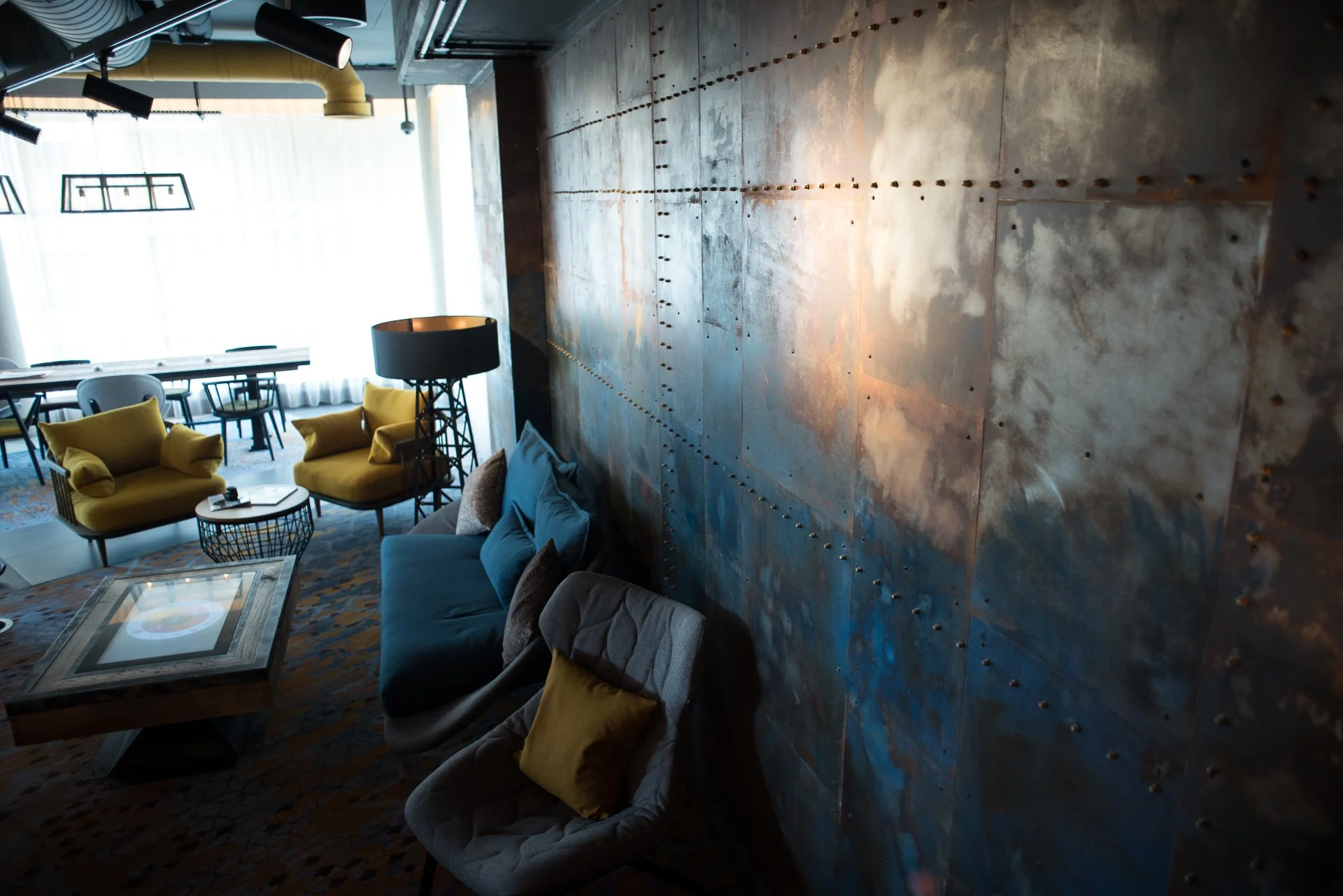 Interior of a modern living room with industrial design, featuring a large, rusted metal wall with rivets, yellow sofas, a glass coffee table, and a seating area with colorful pillows. There are large windows with sheer curtains and black ceiling-mou