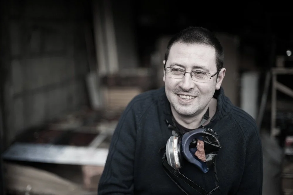 Man with glasses smiling, wearing a dark sweater and safety mask around his neck, in a workshop or industrial setting.