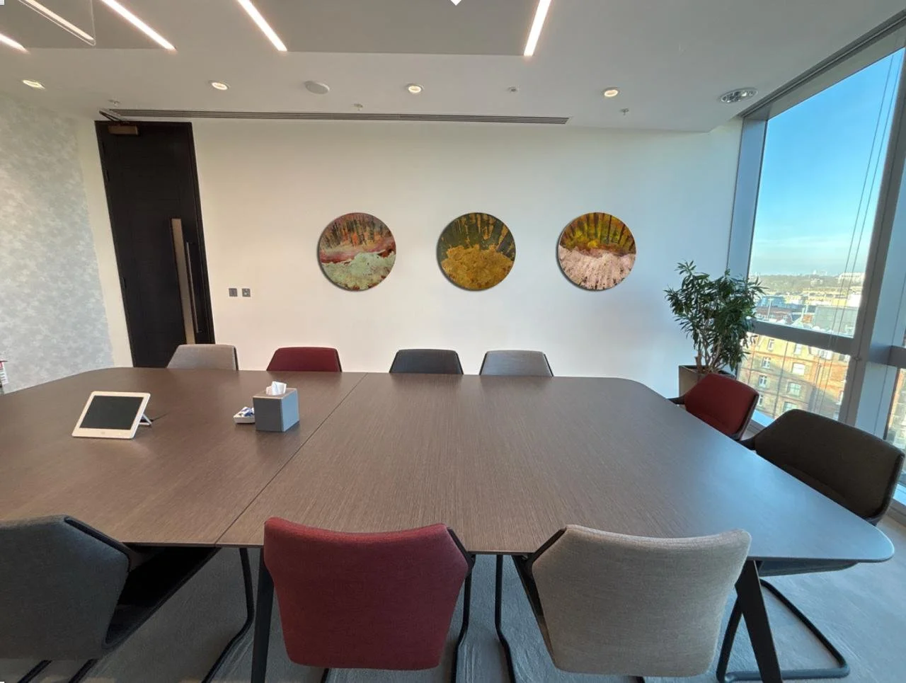 An empty modern conference room with a large oval wooden table, colorful chairs, a tablet, tissue box, and a plant near floor-to-ceiling windows showing cityscape views. Artworks hang on the wall.