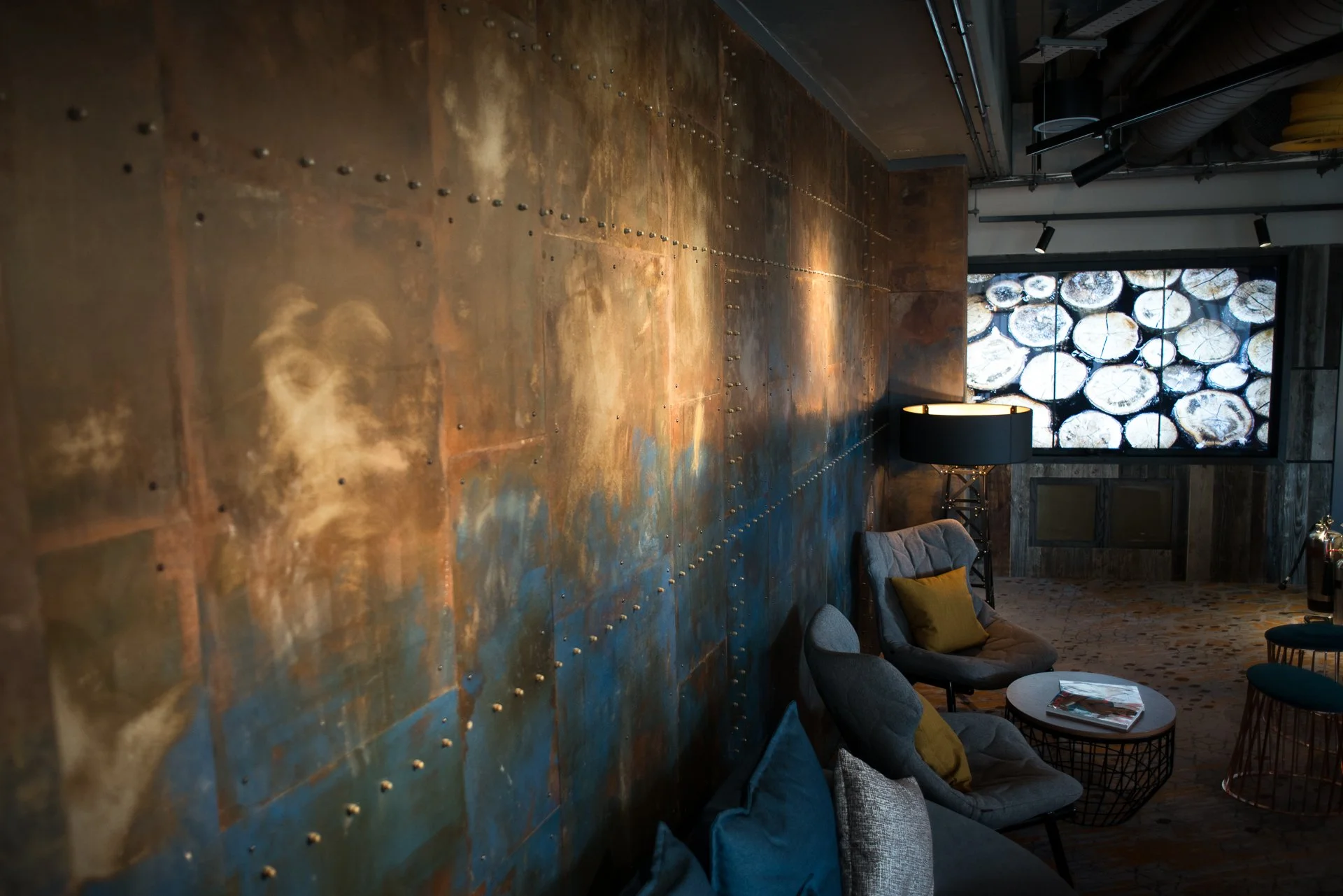 A cozy lounge area with armchairs and a side table with magazines. A large abstract metal wall art with rivets decorates the wall, and there is a window with a decorative window film of logs. A lamp and some exposed ductwork are also visible.