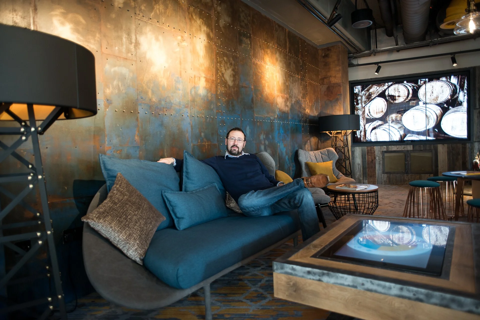 Man sitting on a blue sofa with multiple cushions in a modern lounge area, with textured metallic and rustic wooden wall behind him, and a large digital display screen showing barrel images in a bar or cafe setting.