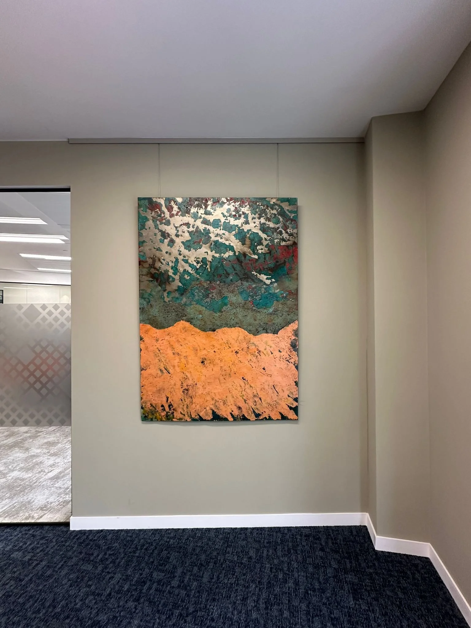 Abstract landscape painting hanging on a plain beige wall in an art gallery with a dark textured carpeted floor.