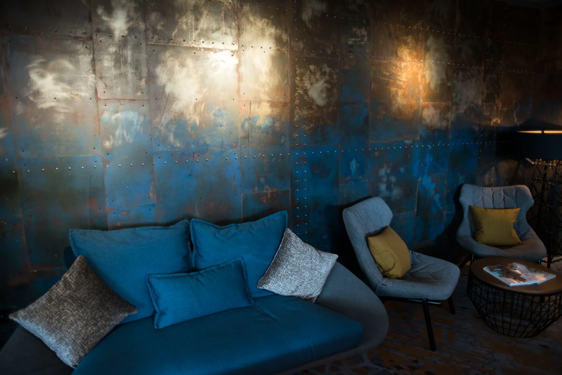 A cozy indoor seating area with a blue sofa and armchairs, dusted with throw pillows in shades of blue, brown, and yellow, set against a rustic, metallic, riveted wall background, with a small round table holding magazines and a stack of books.