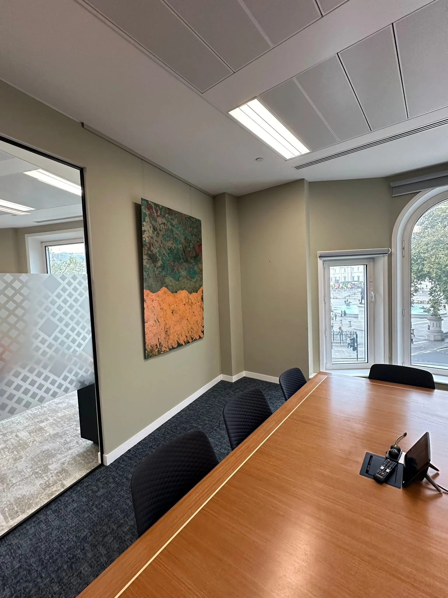 A conference room with a wooden table, black chairs, a window, and a door with a frosted glass panel. There is a painting on the wall, ceiling lights, and a window overlooking city buildings and a street.