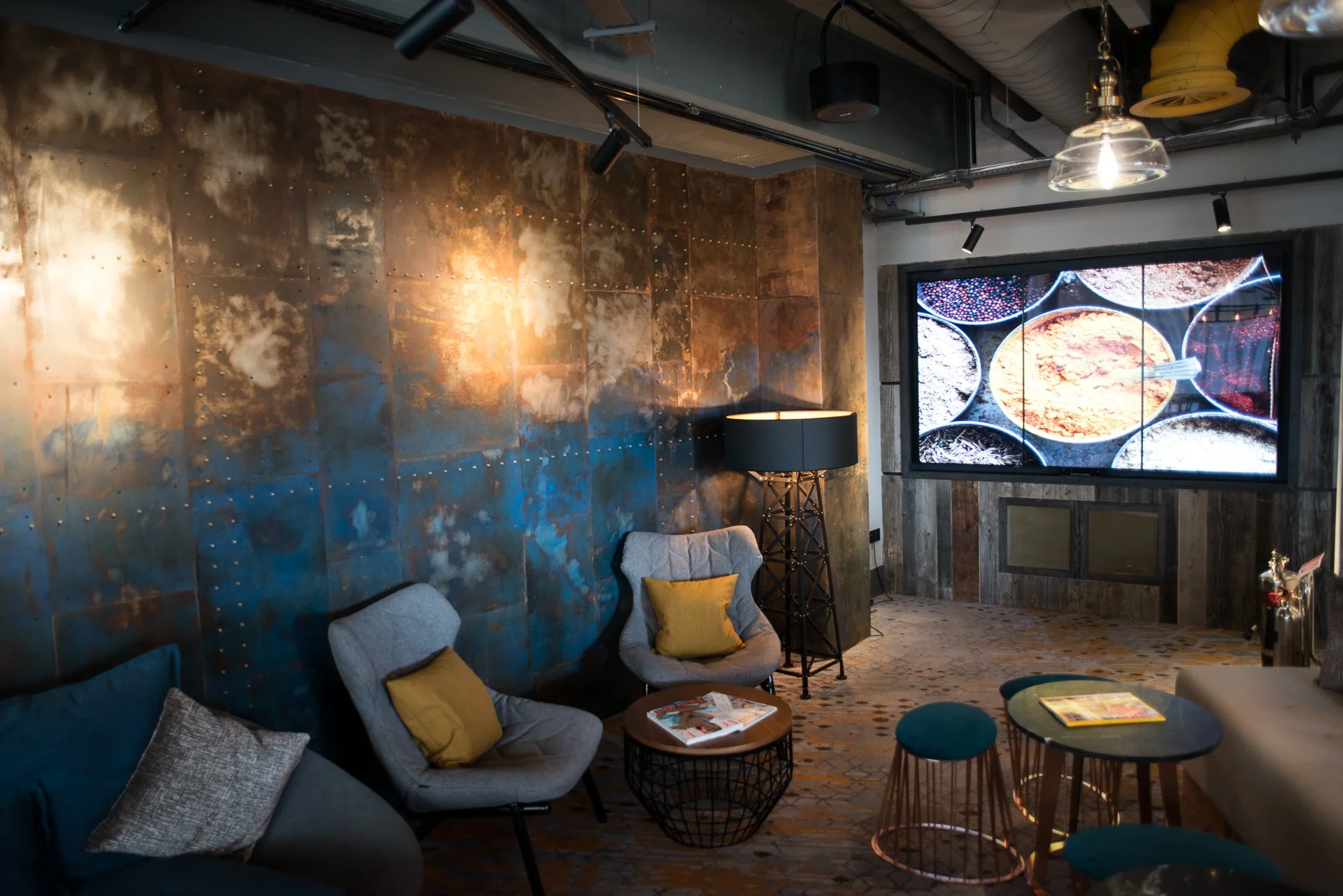 Interior of a modern lounge with a rust-colored metal wall, a large digital screen displaying spices, a black lamp, and comfortable chairs with yellow pillows.