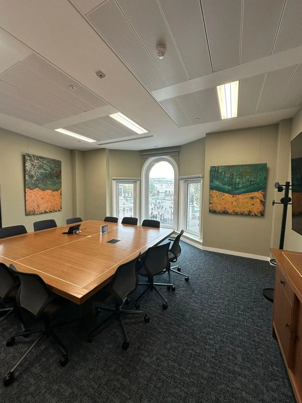 Empty conference room with a large wooden table, black office chairs, two paintings on the walls, large windows showing a cityscape, and a wooden cabinet.