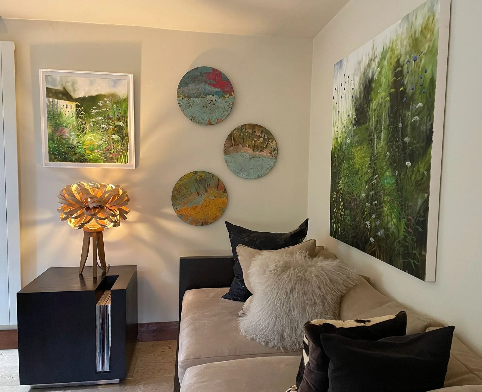 A cozy living room corner with a beige sofa decorated with black, beige, and fuzzy pillows, a wooden side table with a unique lamp, and artwork on the wall including landscape paintings and an abstract floral piece.