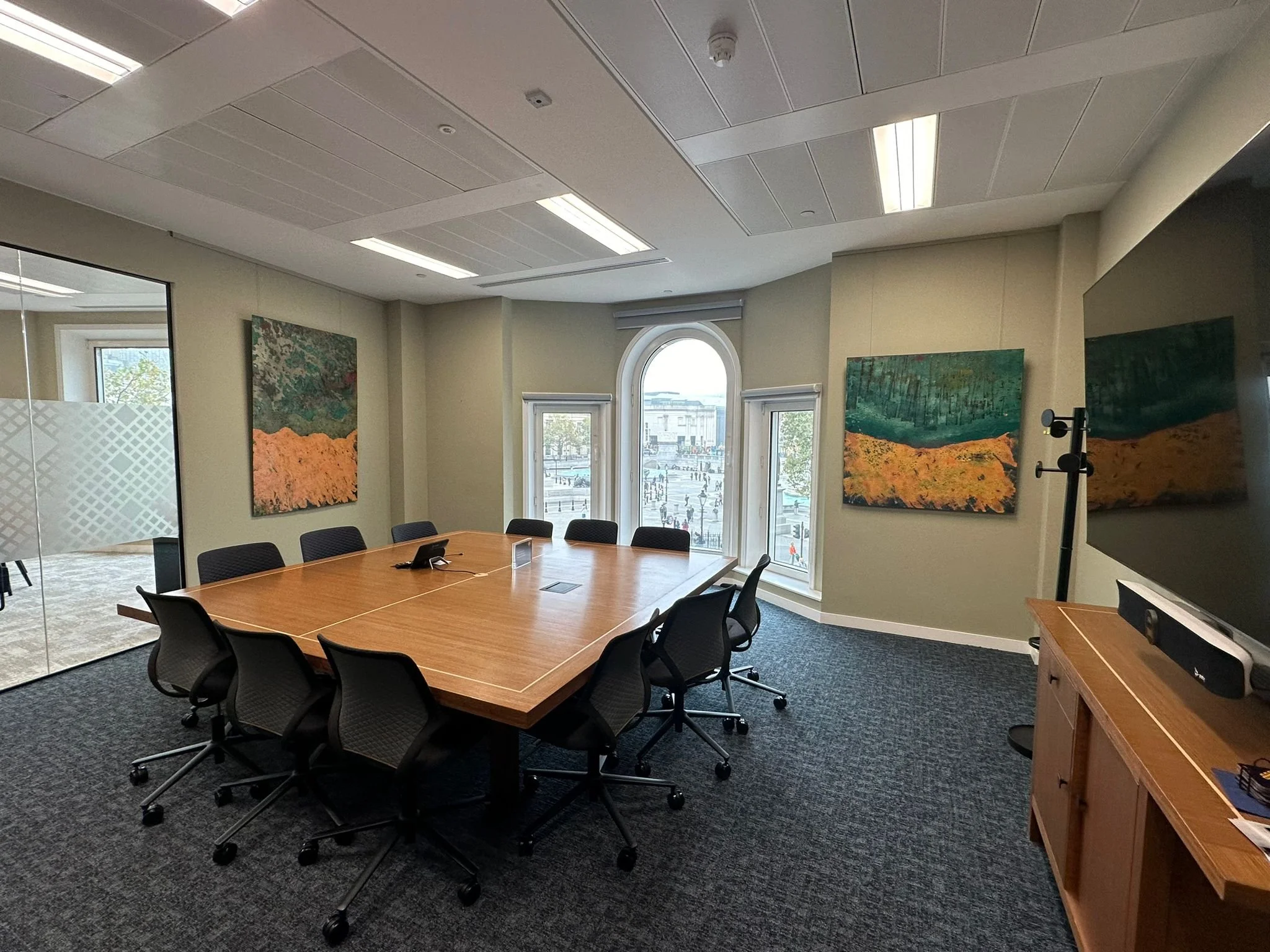 A conference room with a rectangular wooden table surrounded by black office chairs on a dark carpeted floor. There are paintings on the walls, large windows with a city view, and a television on the right side of the image.