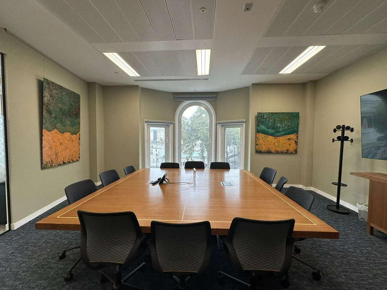 Empty conference room with a large wooden table, black chairs, a coat rack, and artwork on the walls, with a large window at the back letting in natural light.