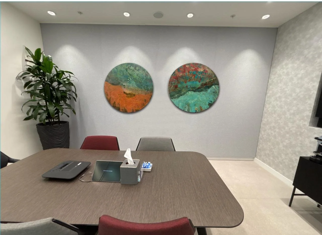 A conference room with a wooden table, four chairs (two red, one gray, one without visible color), a tissue box, a tablet, and a conference phone on the table. There is a large potted plant in a black textured pot on the left side, and two circular a