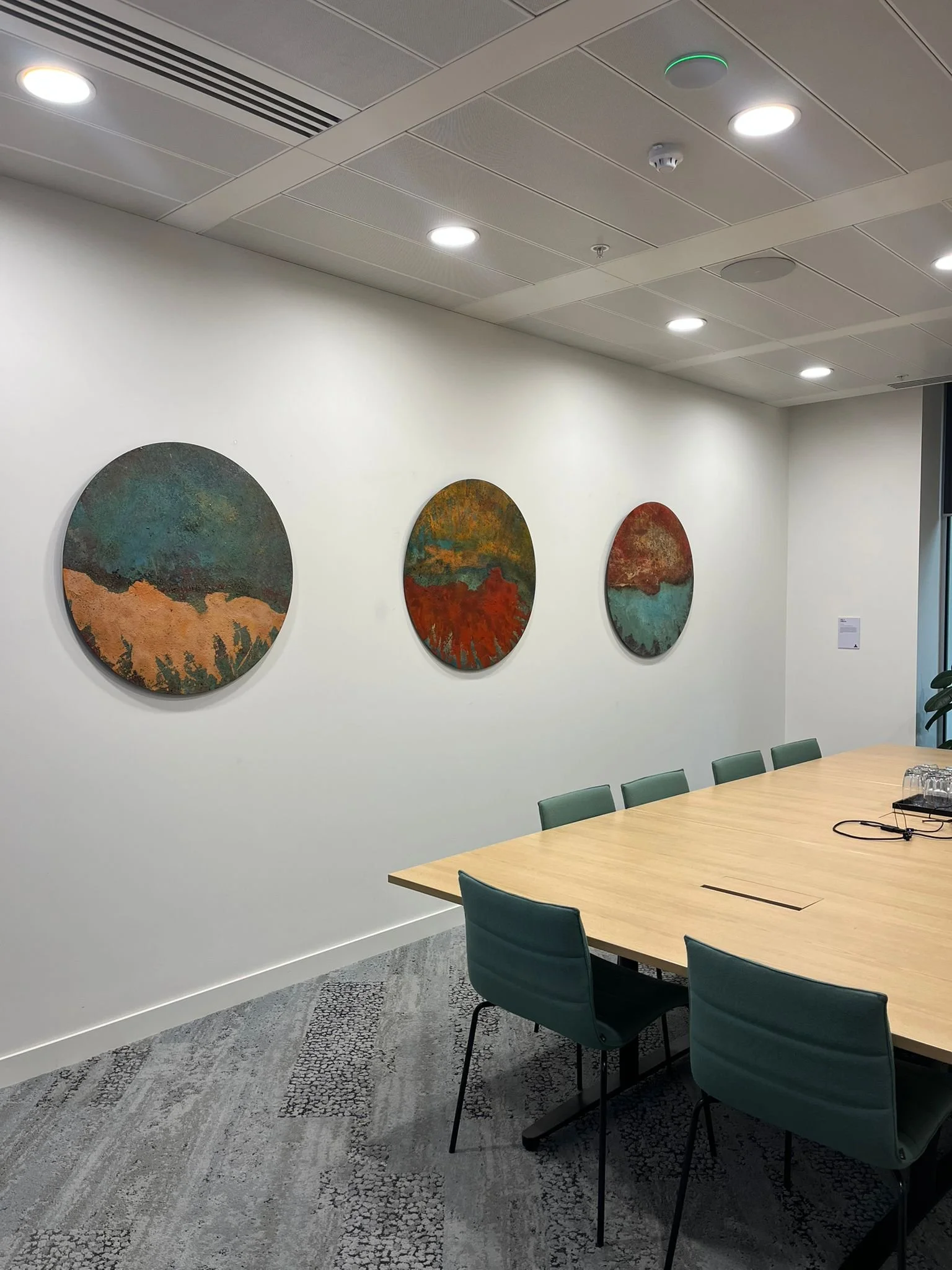 An interior conference room with a long wooden table, teal chairs, and three circular abstract paintings on a white wall. The ceiling has recessed lighting, vents, and a smoke detector.