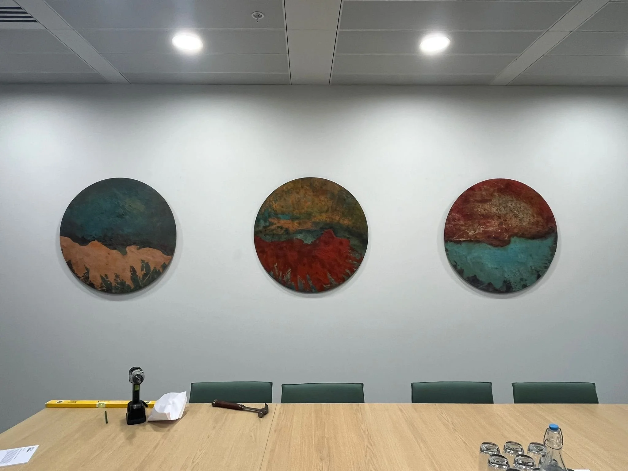 Conference room with a large wooden table, three green chairs, wall art of three circular landscape paintings, a white wall, ceiling lights, and a few items on the table including a tissue box, glasses, and a small blue item.