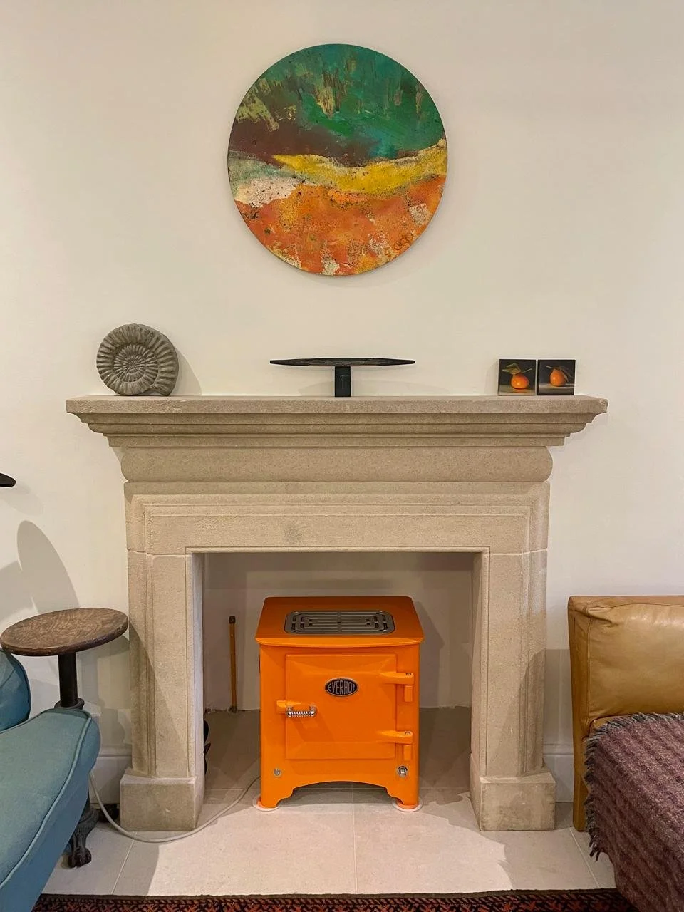 Living room with a beige fireplace mantel decorated with a gray ammonite fossil, a black tray, and two small paintings of gourds. Above the mantel, there is an abstract round painting with green, yellow, white, orange, and brown colors. Inside the fi