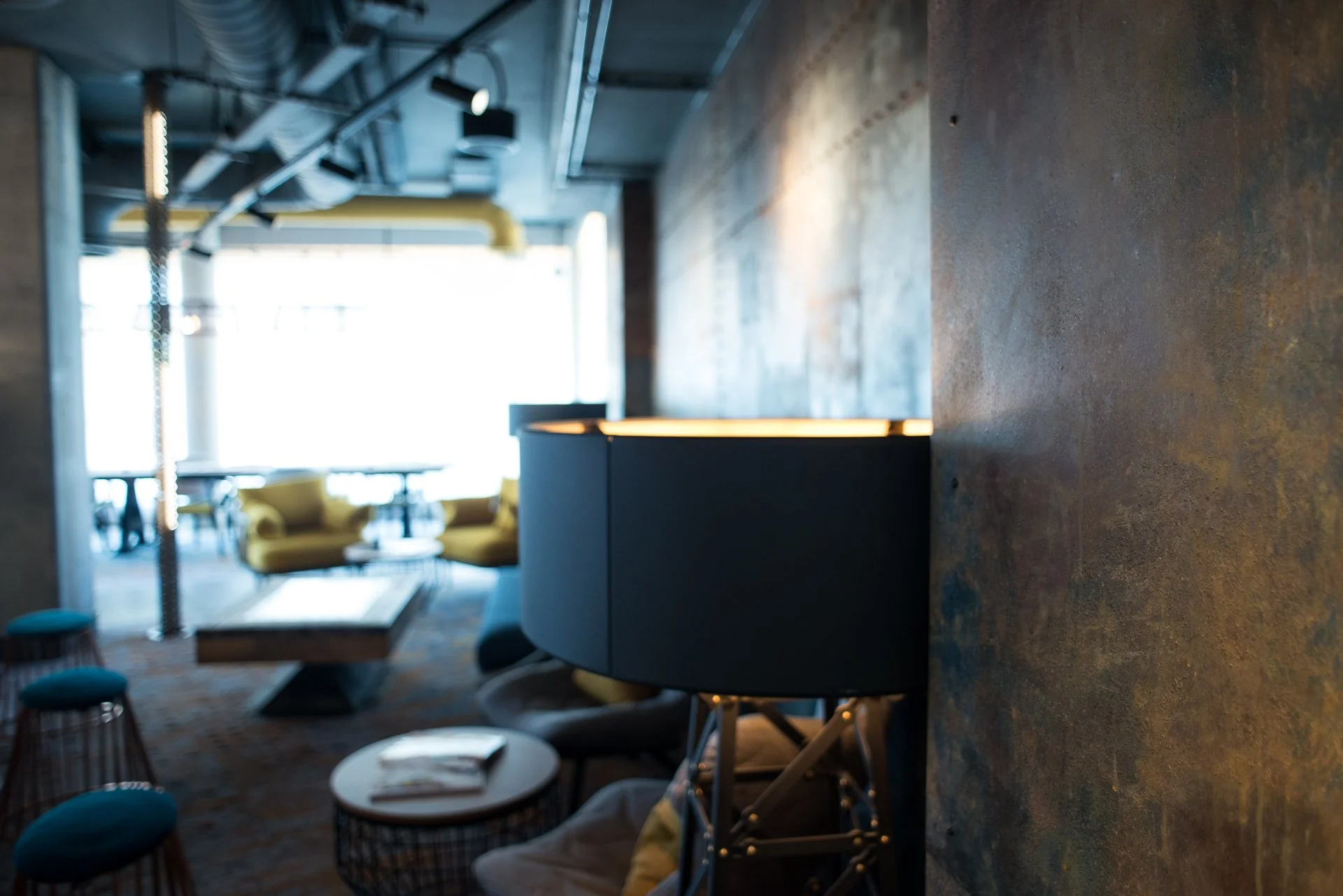 Modern indoor space with bar seating, yellow chairs, and large windows providing natural light, industrial ceilings with exposed ductwork and lighting fixtures.