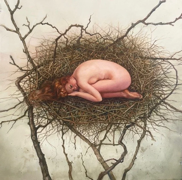 A woman with red hair curled up on her side inside a nest made of twigs, looking at the camera.