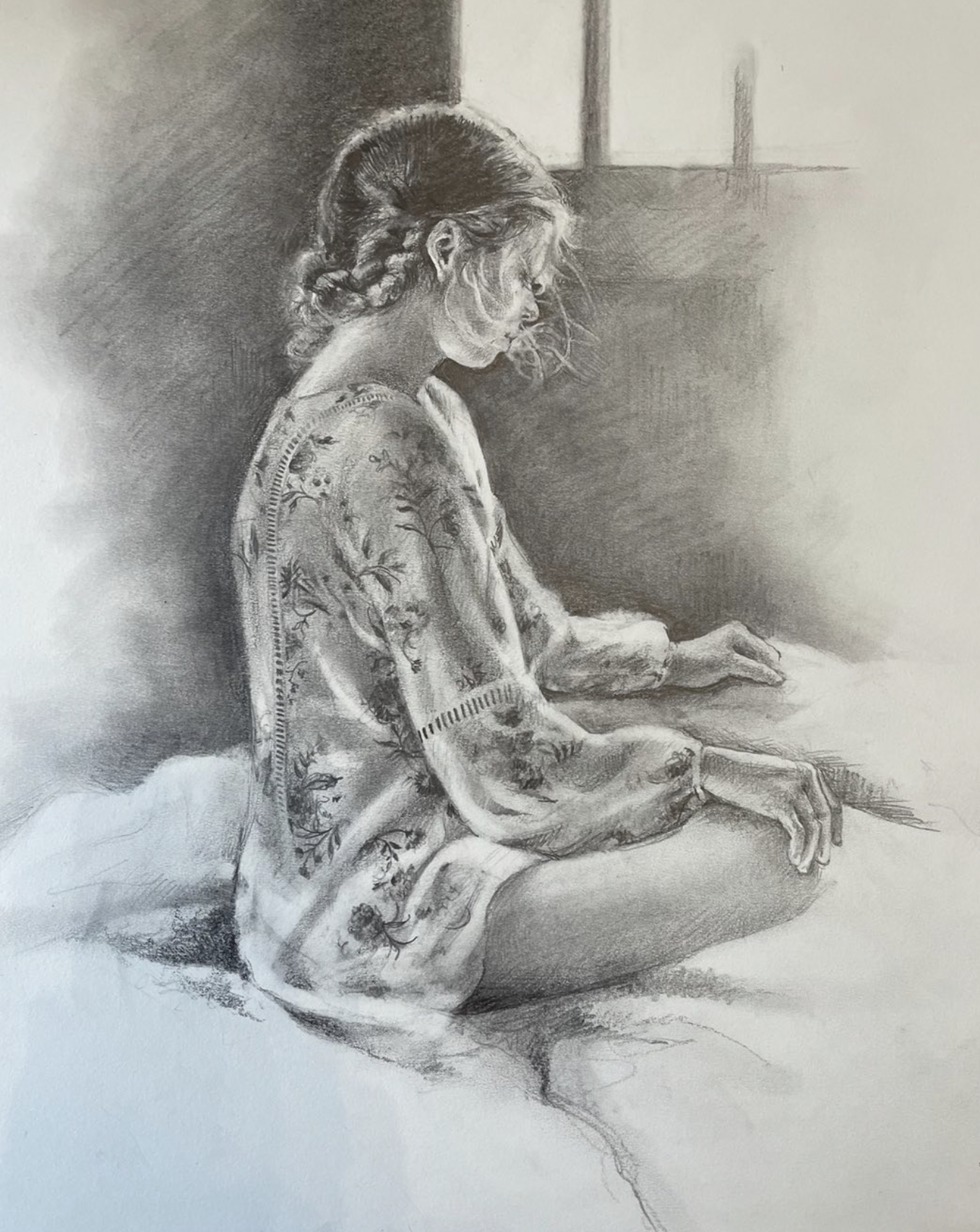 A detailed pencil drawing of an elderly woman with glasses, seated and looking towards her right, with her hands resting on her lap. She has her hair tied back and is dressed in a patterned, long-sleeved garment. The background is simple with faint l