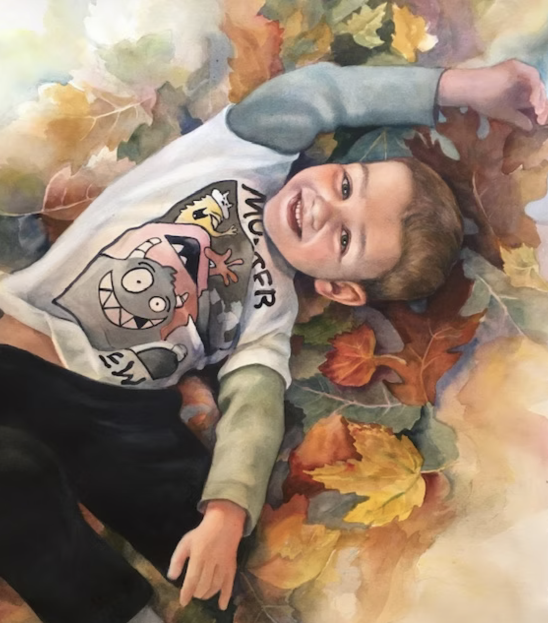 A smiling boy lying on a bed of colorful autumn leaves wearing a white sweatshirt with cartoon characters.