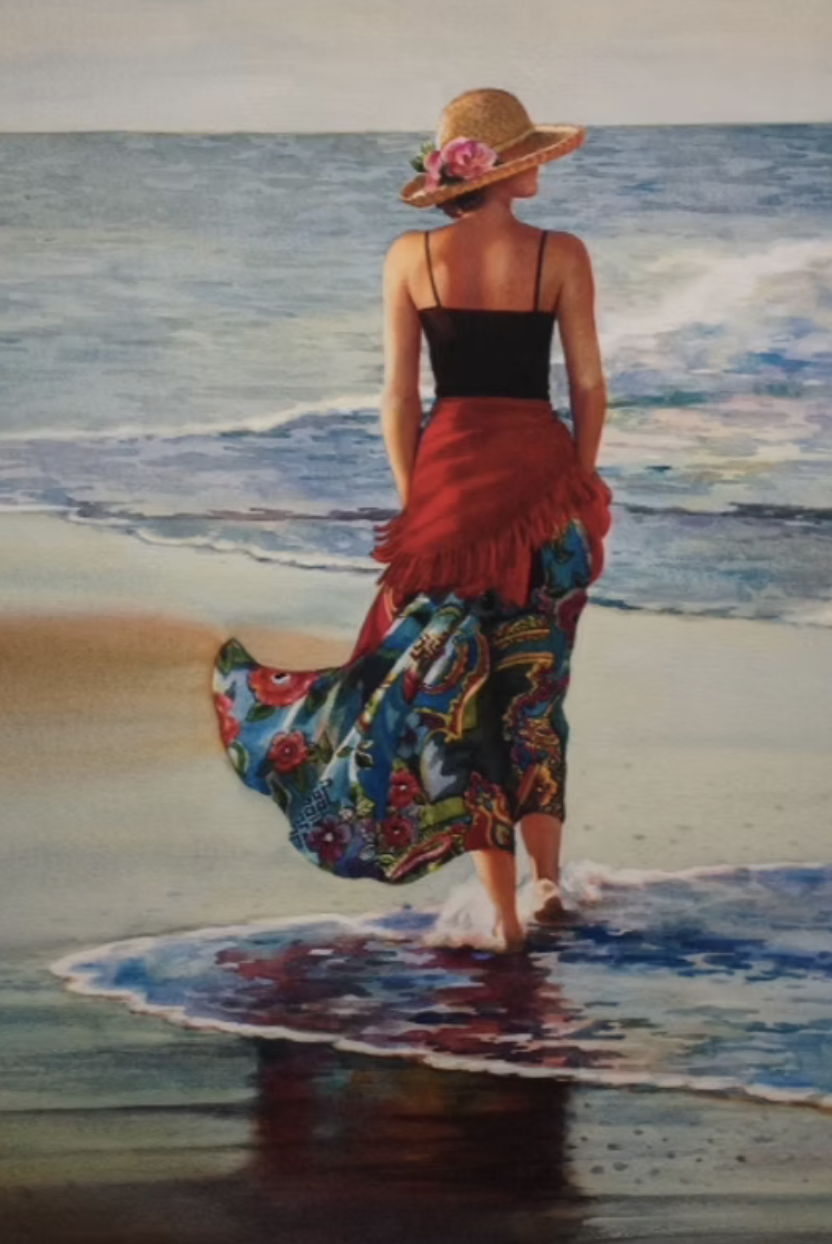 Woman in a black top and red shawl walking along the shoreline of the beach, with a colorful scarf wrapped around her waist and a sunhat decorated with pink flowers.