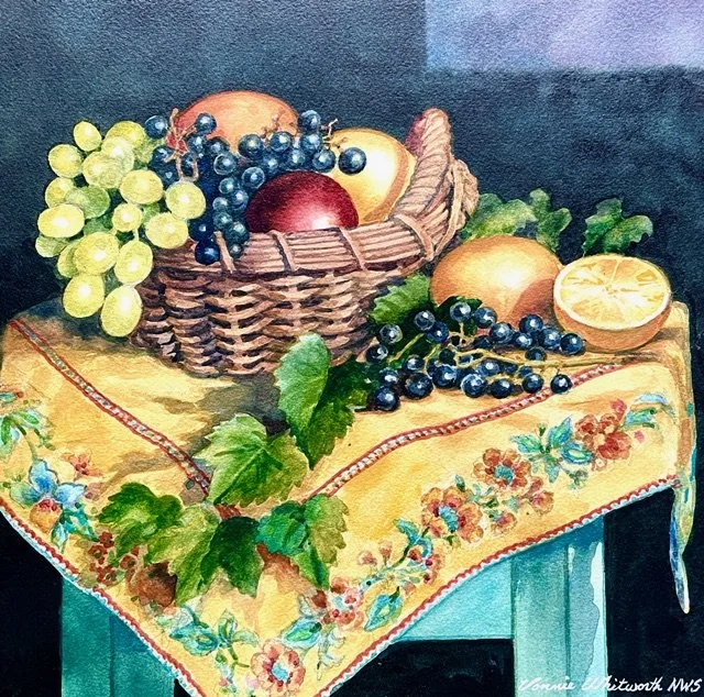 A basket of grapes and peaches on a table with a decorative tablecloth, beside a lemon half and green leaves.