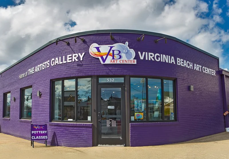 Exterior of the Virginia Beach Art Center, a purple building with large front windows and a sign indicating pottery classes. The sky is partly cloudy.