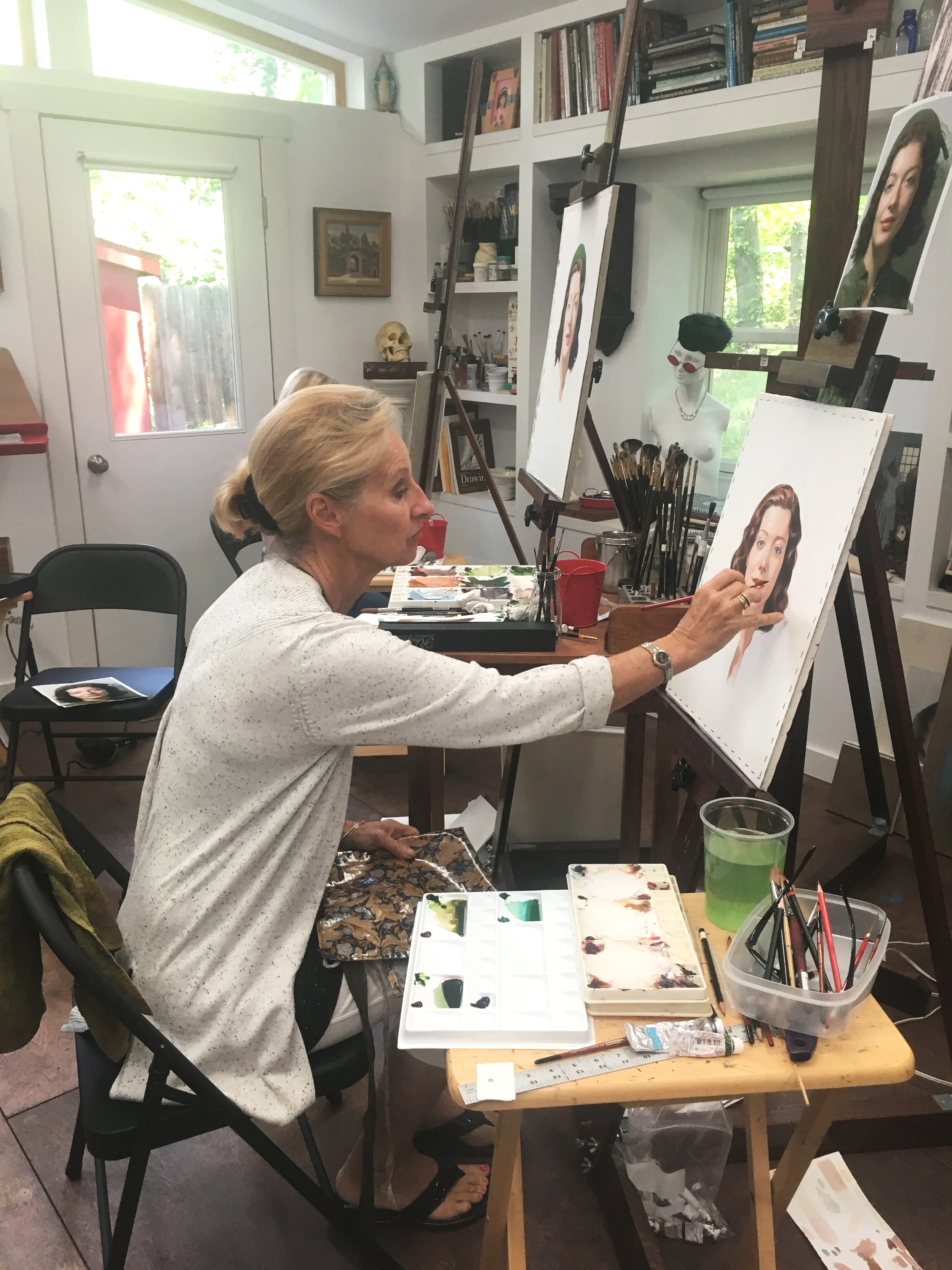 An elderly woman is painting a realistic portrait of a woman with dark hair and a green shirt on an easel in an art studio. She is sitting at a wooden table with a palette, brushes, and paint supplies. The room has shelves with books and art materials, a window, and some artwork on the walls.