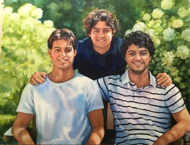 Three young men sitting outdoors in front of lush green foliage, smiling at the camera, with the man in the middle standing behind and placing his hands on the shoulders of the other two.