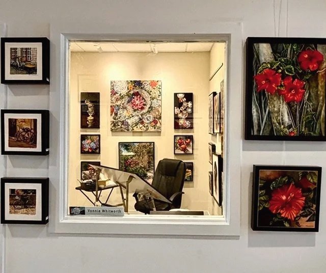 View through a window showing an art gallery with paintings on the walls, including floral and scenic artwork, a desk with a chair, and a nameplate that reads "Vonda Whitehurst."