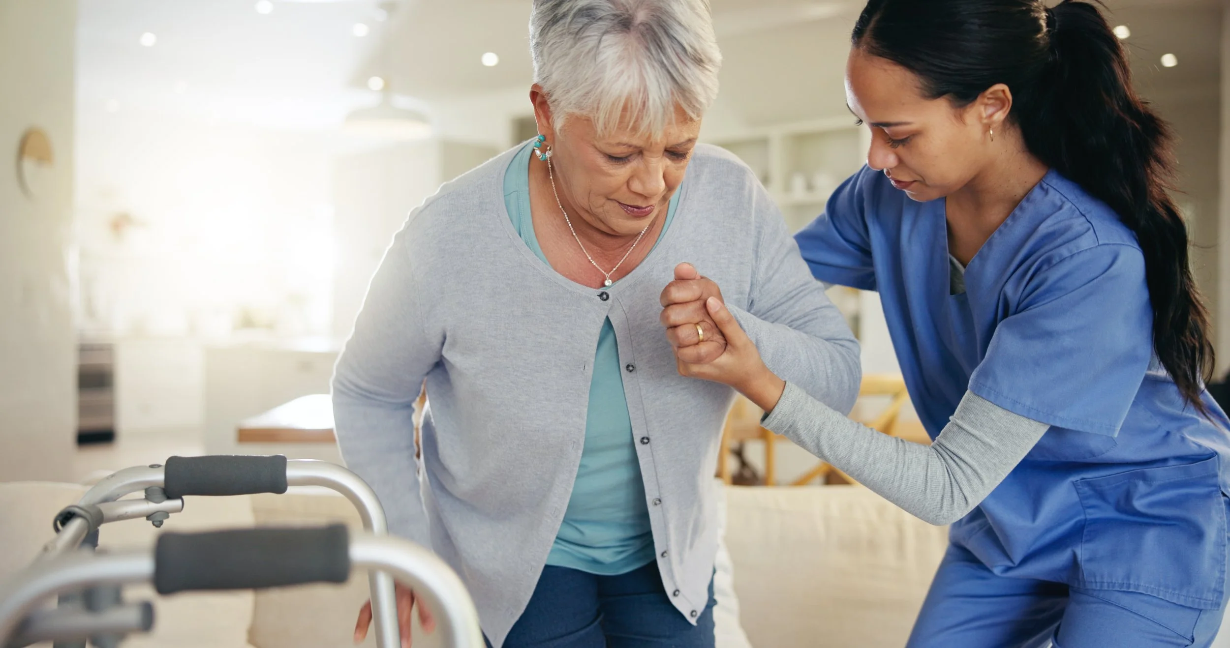 A young female nurse assisting an elderly woman with a walker in a healthcare setting, holding her hand and providing support.