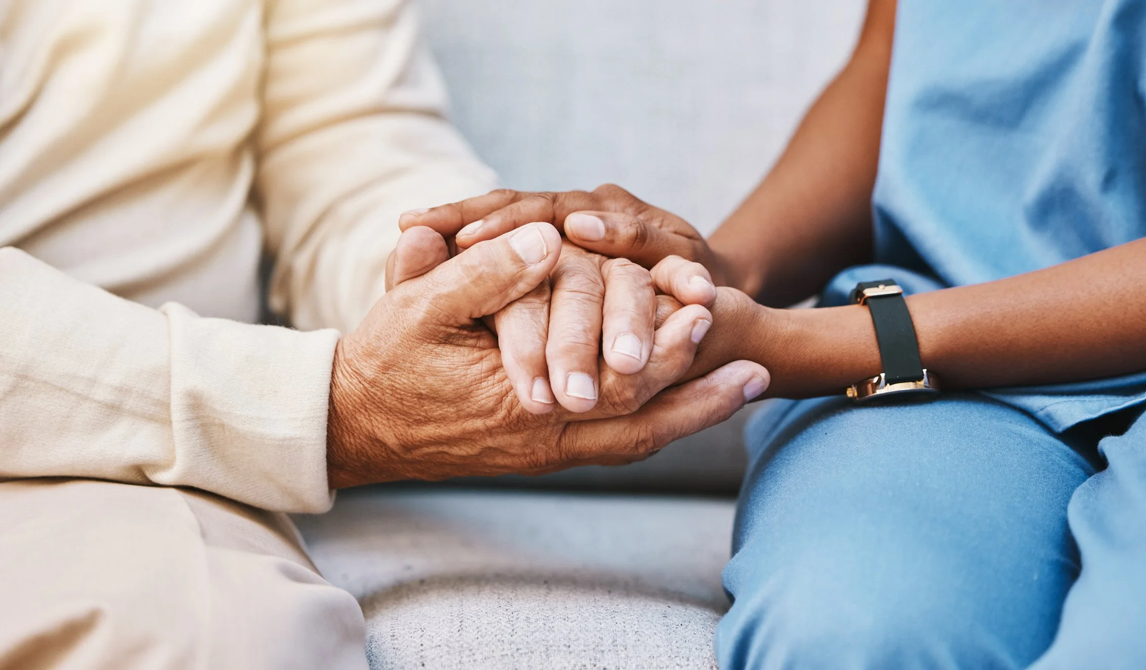 Close-up of two people holding hands, one older with darker skin and a beige sleeve, the other younger with lighter skin, a black wristwatch, and a blue sleeve.